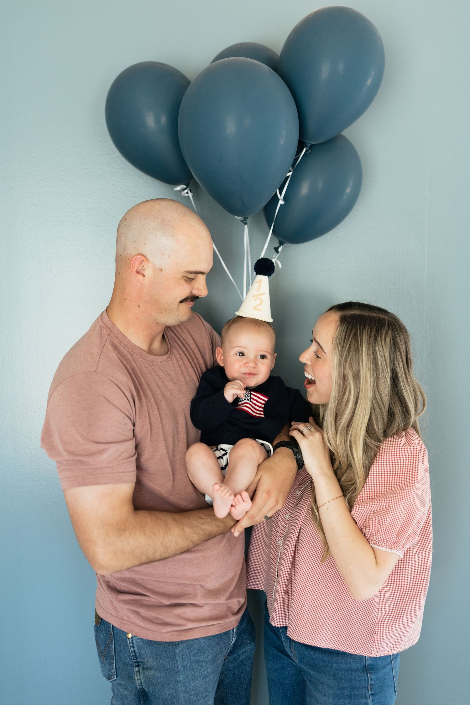 A couple celebrates their baby's first birthday with blue balloons and a party hat against a light blue wall.