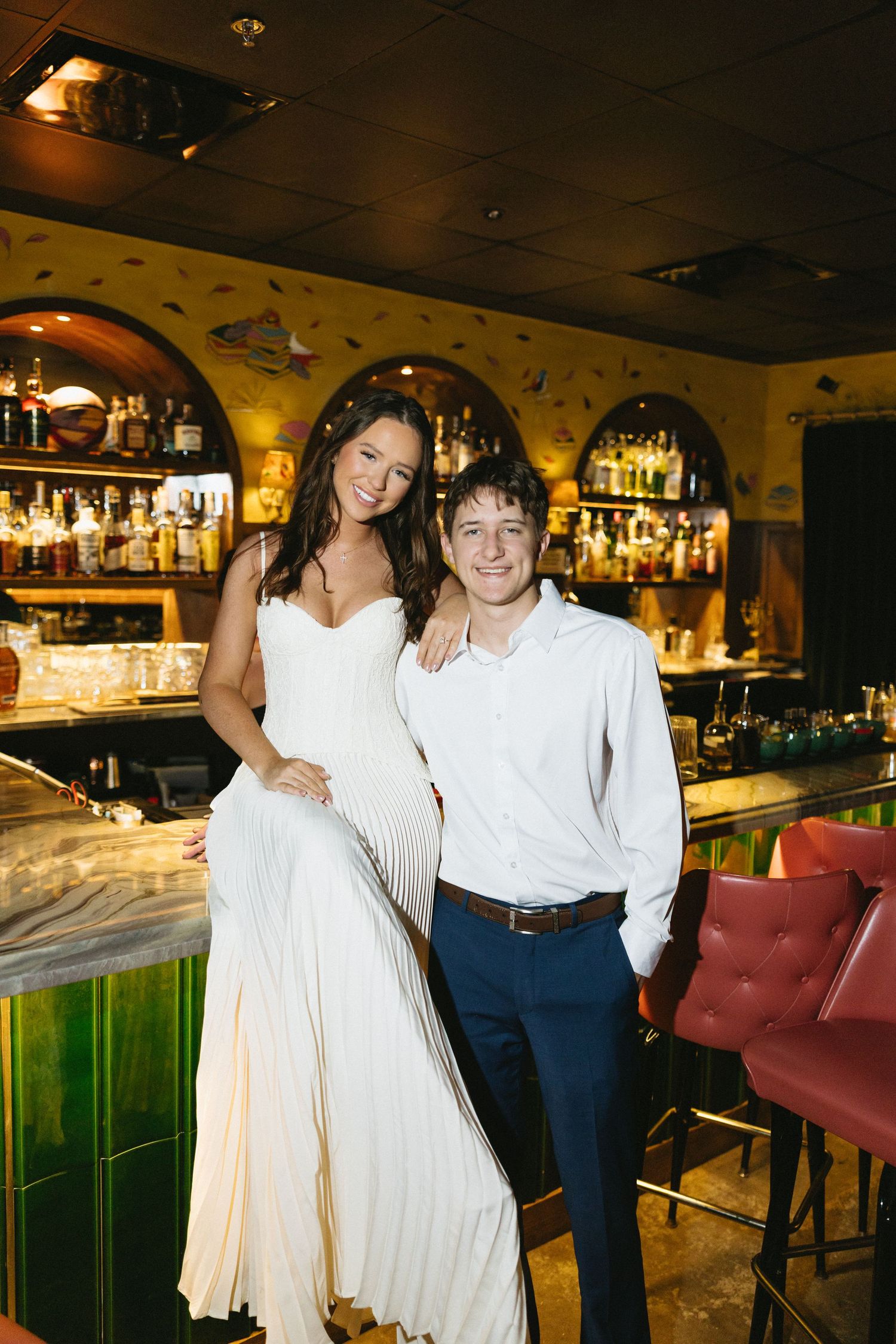 A couple in wedding attire smiles together at a warmly lit bar with arched shelves of liquor bottles behind them.