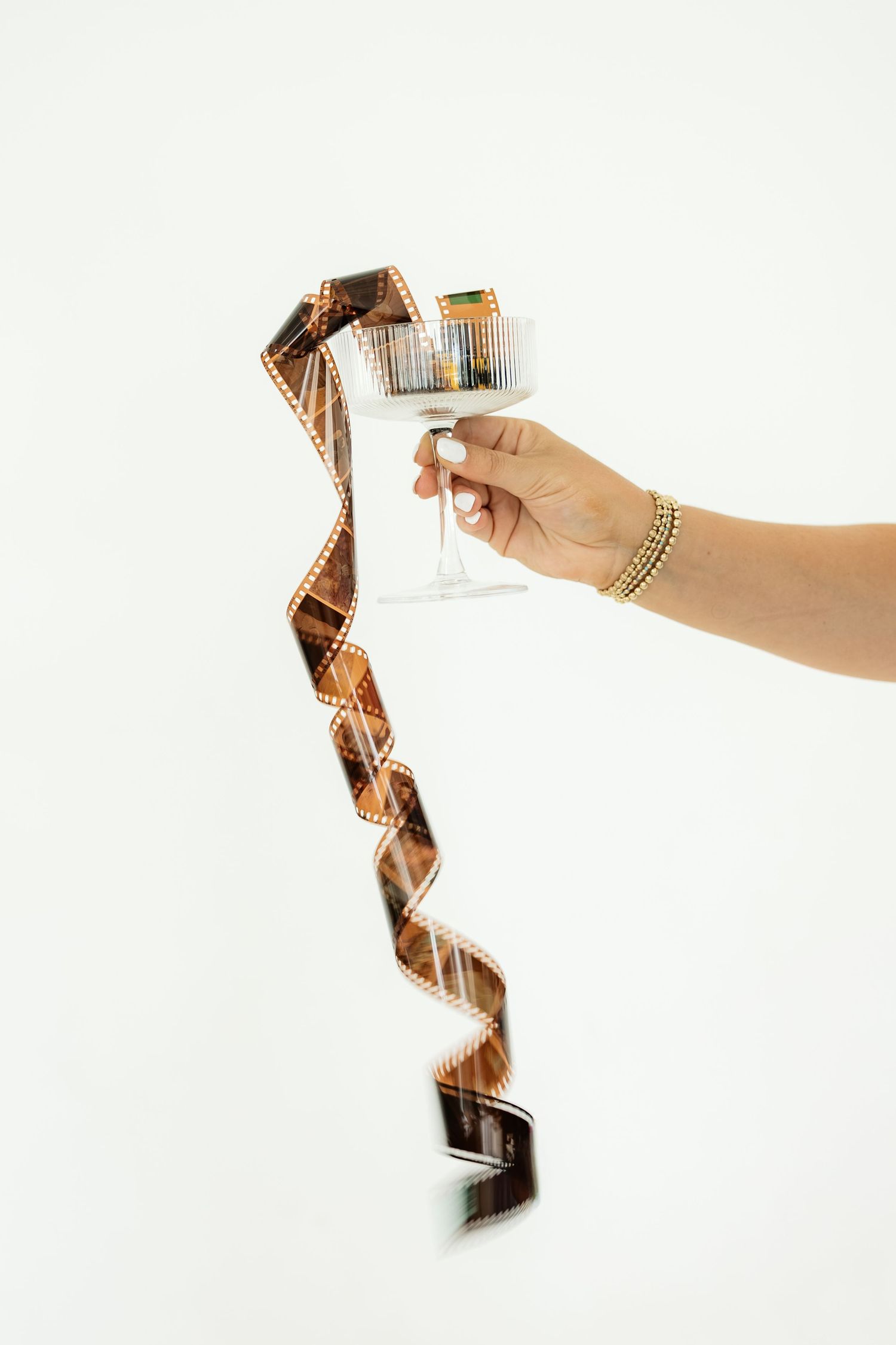 Hand holding a champagne glass filled with spiraling 35mm film strip on white background.