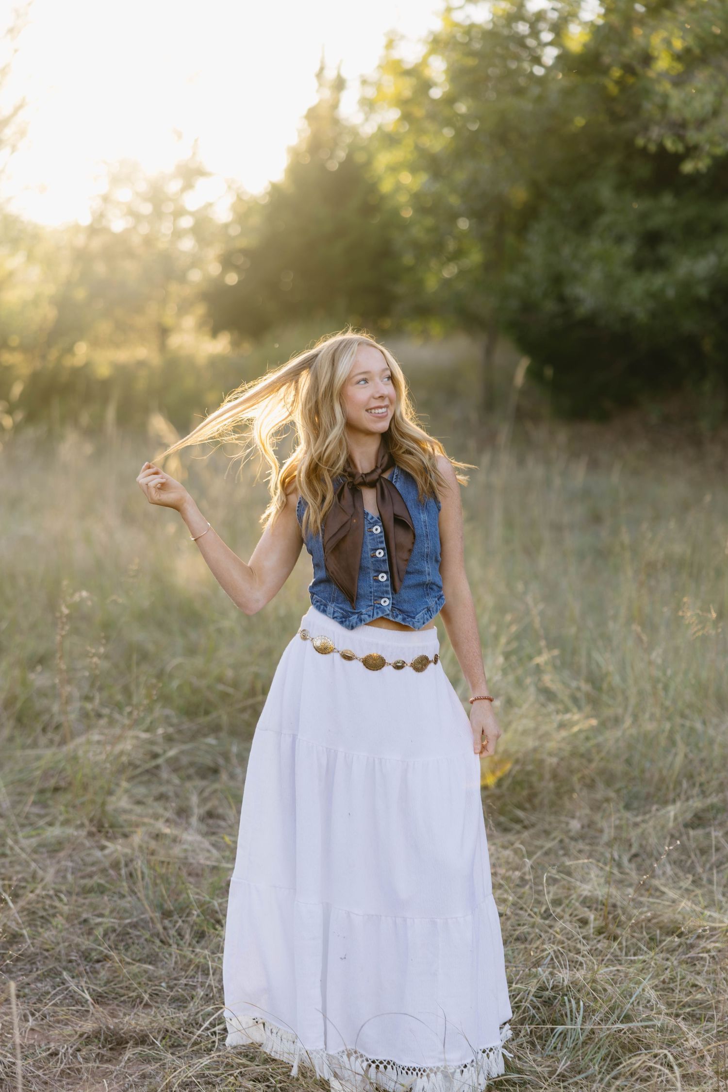 Young blonde woman in denim vest and white maxi skirt twirling her hair in a sunlit field.
