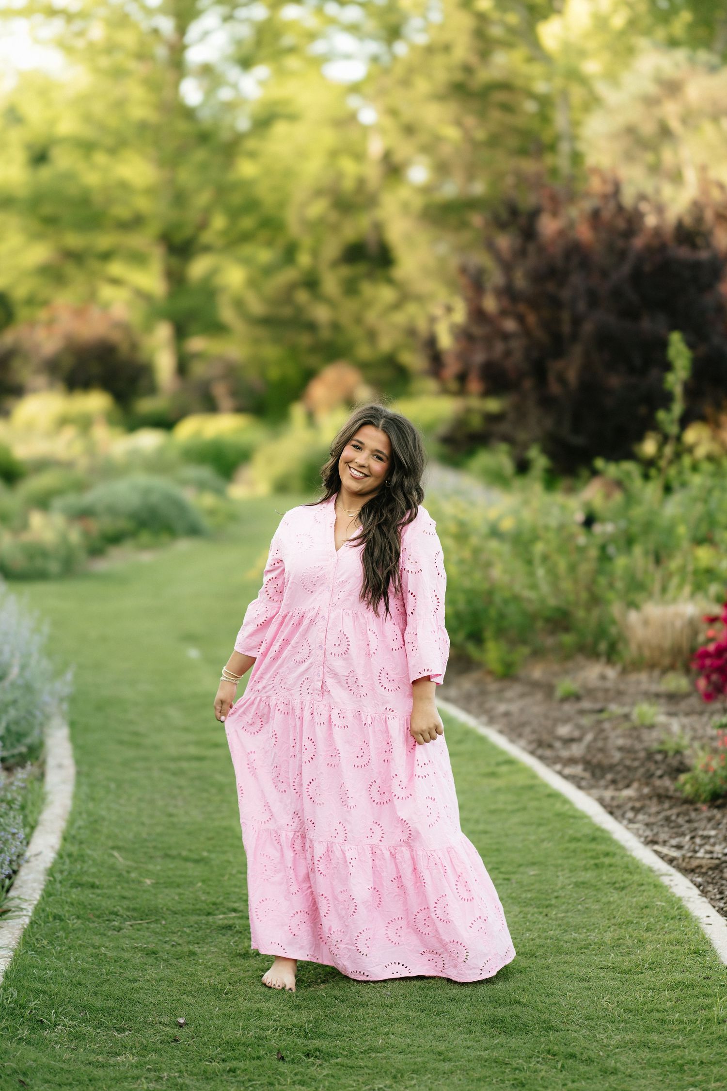 Young woman in pink paisley maxi dress walking barefoot on green garden path surrounded by lush greenery.