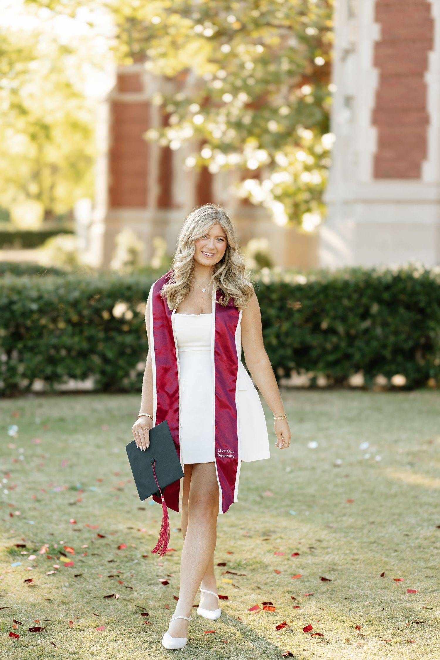 Smiling blonde woman in white dress with maroon graduation stole walking on a college campus.