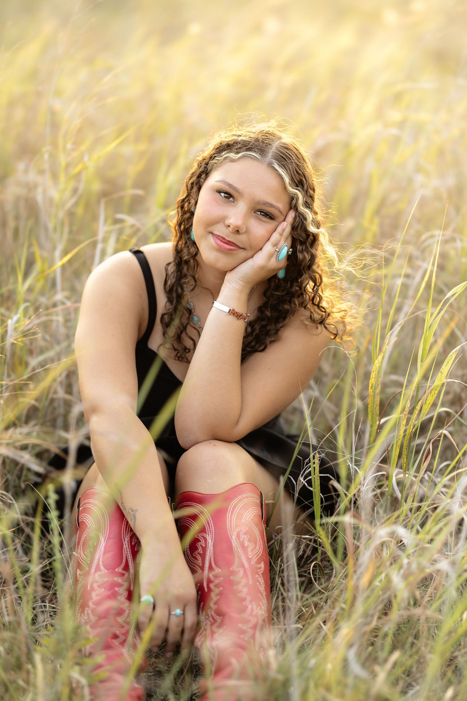 Young woman with curly hair in black top and pink cowboy boots sitting in tall golden grass field.
