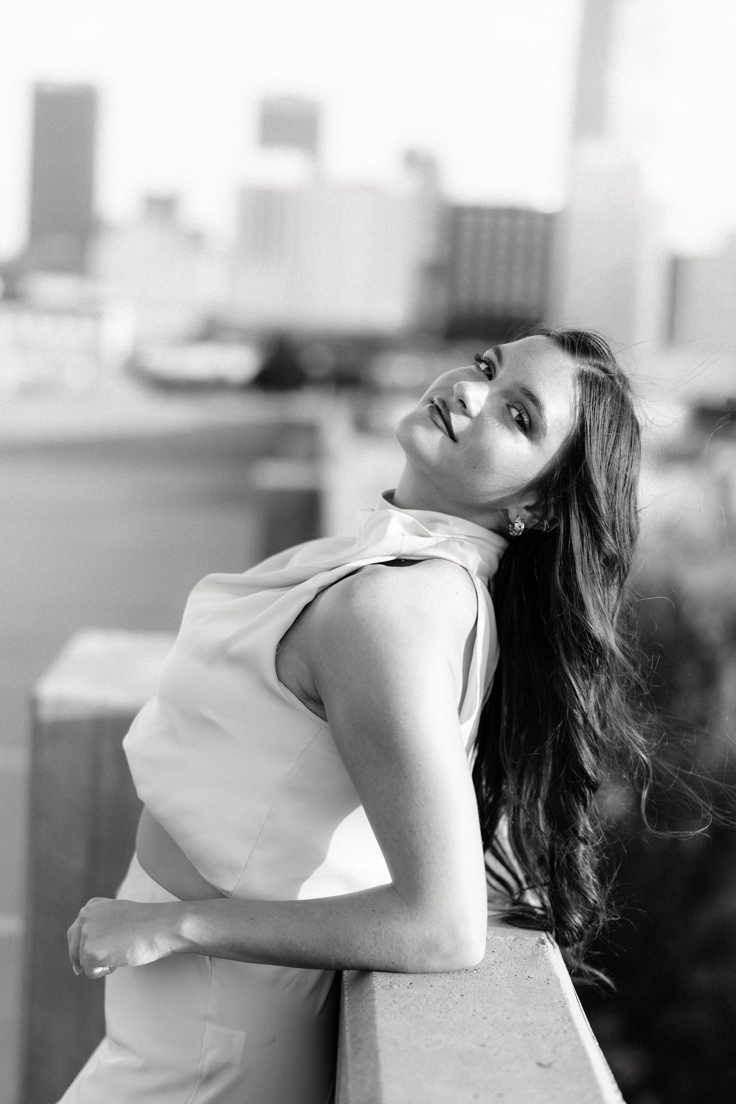 Black and white portrait of a woman with long hair leaning on a railing with a city skyline backdrop.