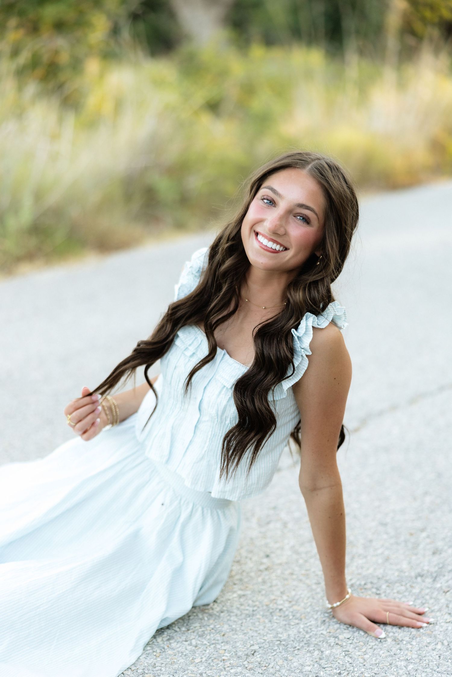 Young woman in white dress sitting on road, smiling, with long wavy brown hair and blue eyes outdoors.