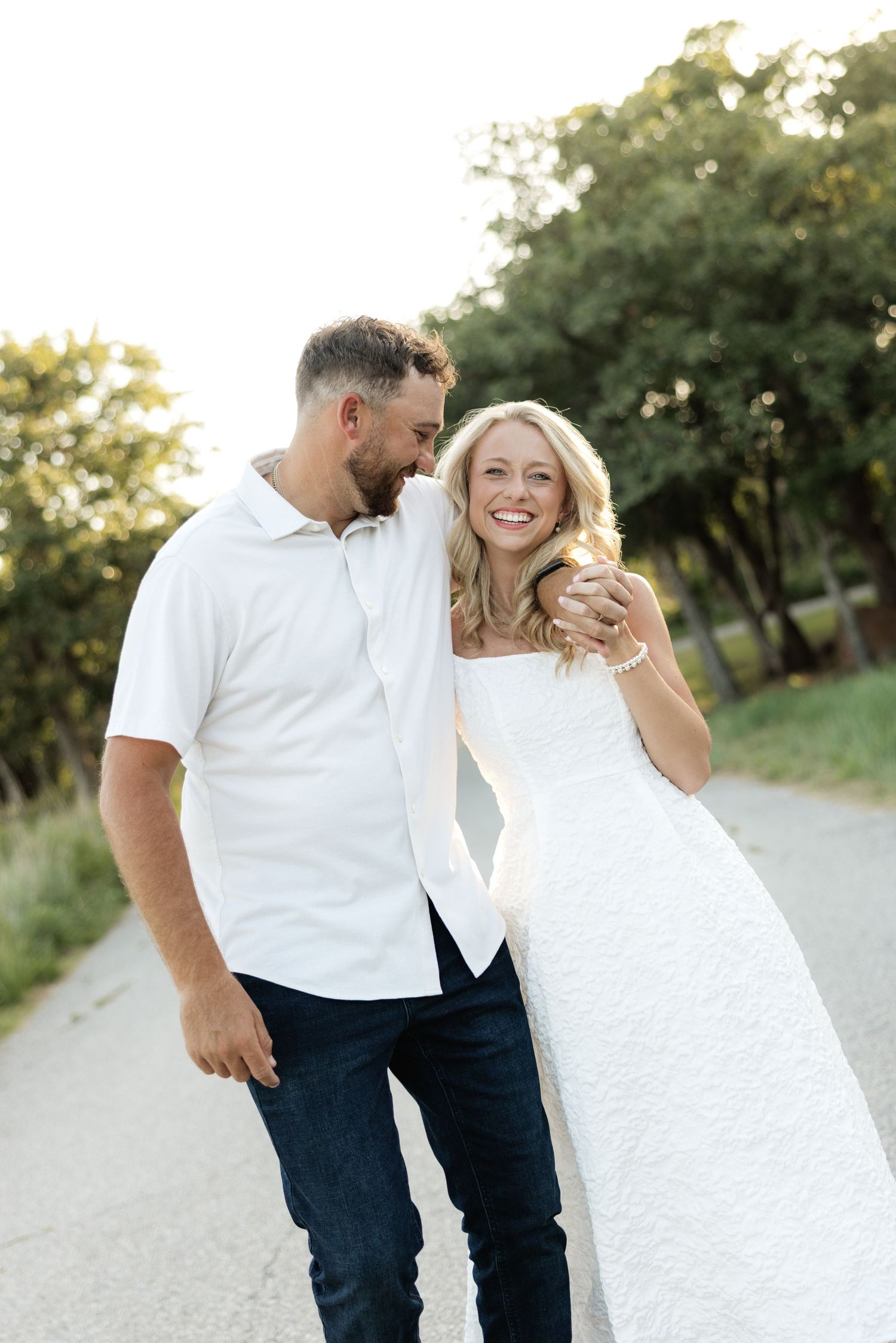 A smiling couple walks outdoors, woman in white dress, man in white shirt and jeans, surrounded by lush green trees.