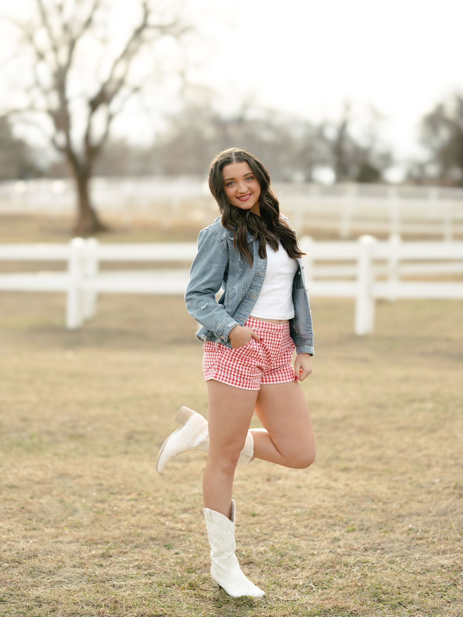 Teen girl in denim jacket pink gingham shorts and white cowboy boots at horse ranch.