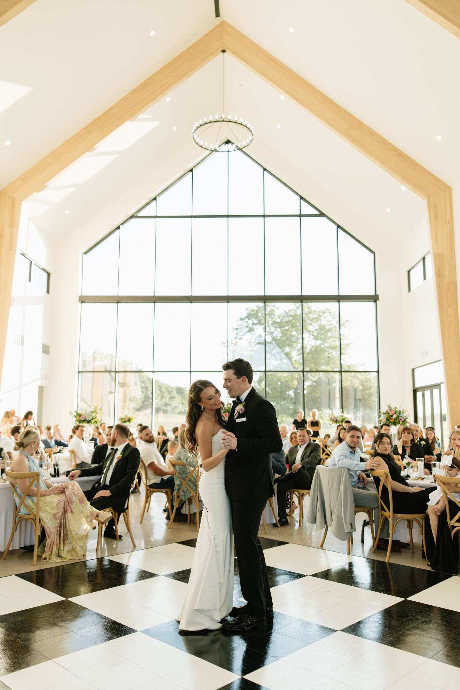 Bride and groom share first dance on checkered floor in stunning light-filled wedding venue.