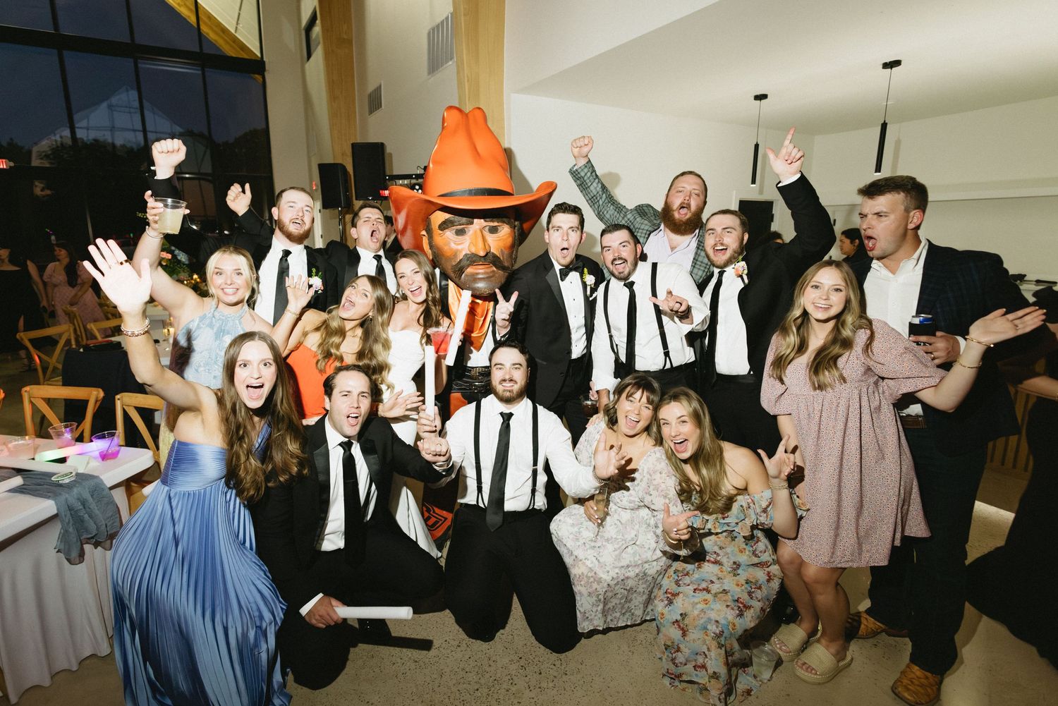 Large wedding party group photo with guests posing enthusiastically with a mascot at the reception.
