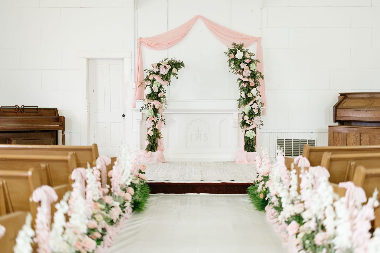 Elegant wedding ceremony arch adorned with pink draping and blush floral arrangements in white chapel.