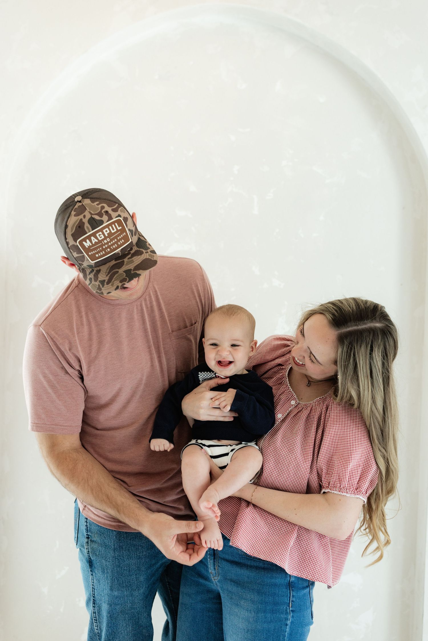 A young couple in casual pink shirts holds their smiling baby boy in front of a white arched wall.