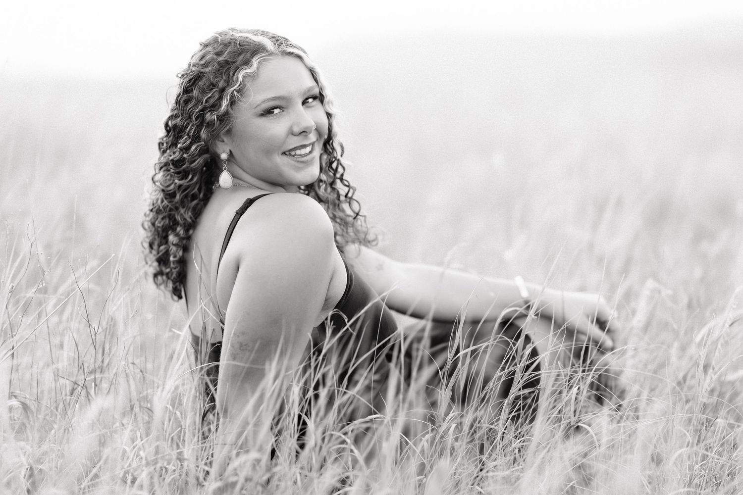 Black and white photo of a smiling young woman with curly hair sitting in a tall grass field.
