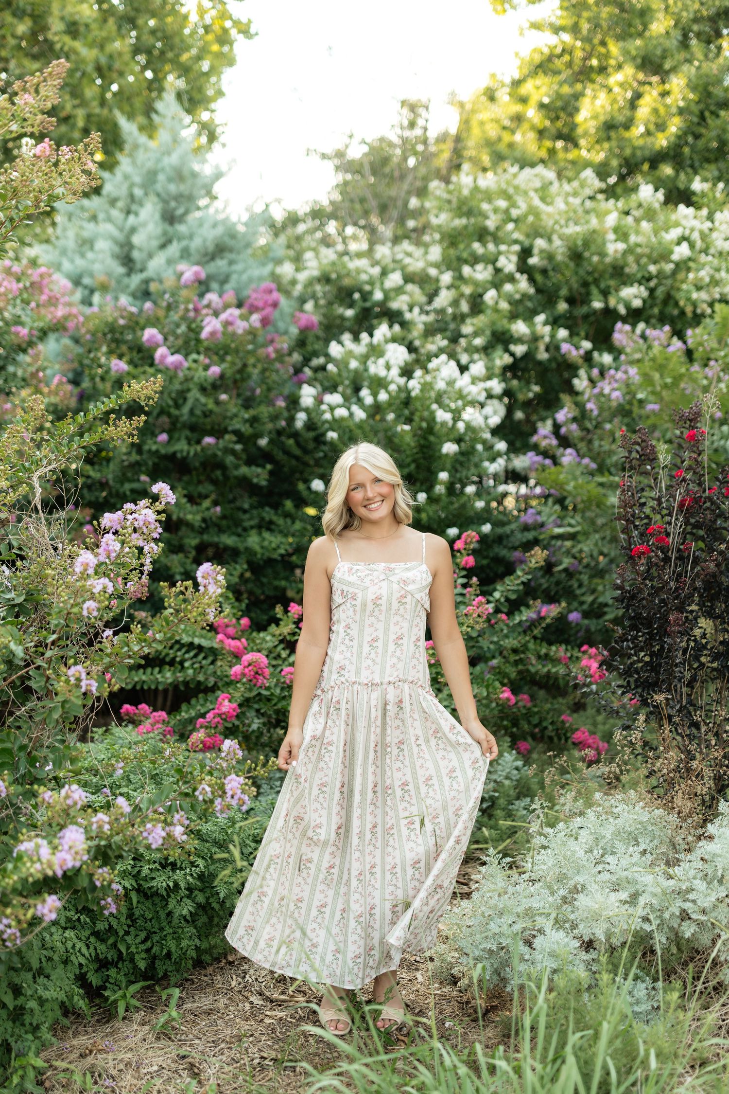 A smiling blonde woman in a floral sundress stands among lush blooming garden bushes with pink and white flowers.