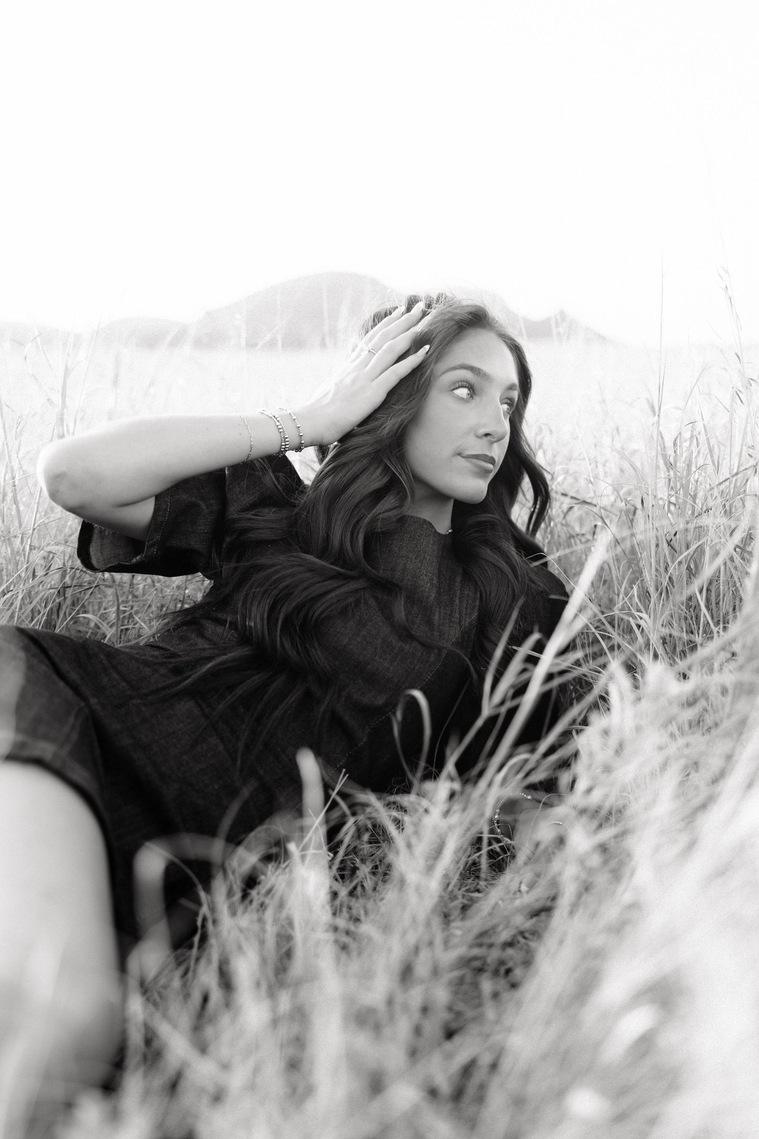 Black and white photo of a woman in a dark dress sitting in tall grass, hand in hair, gazing away.