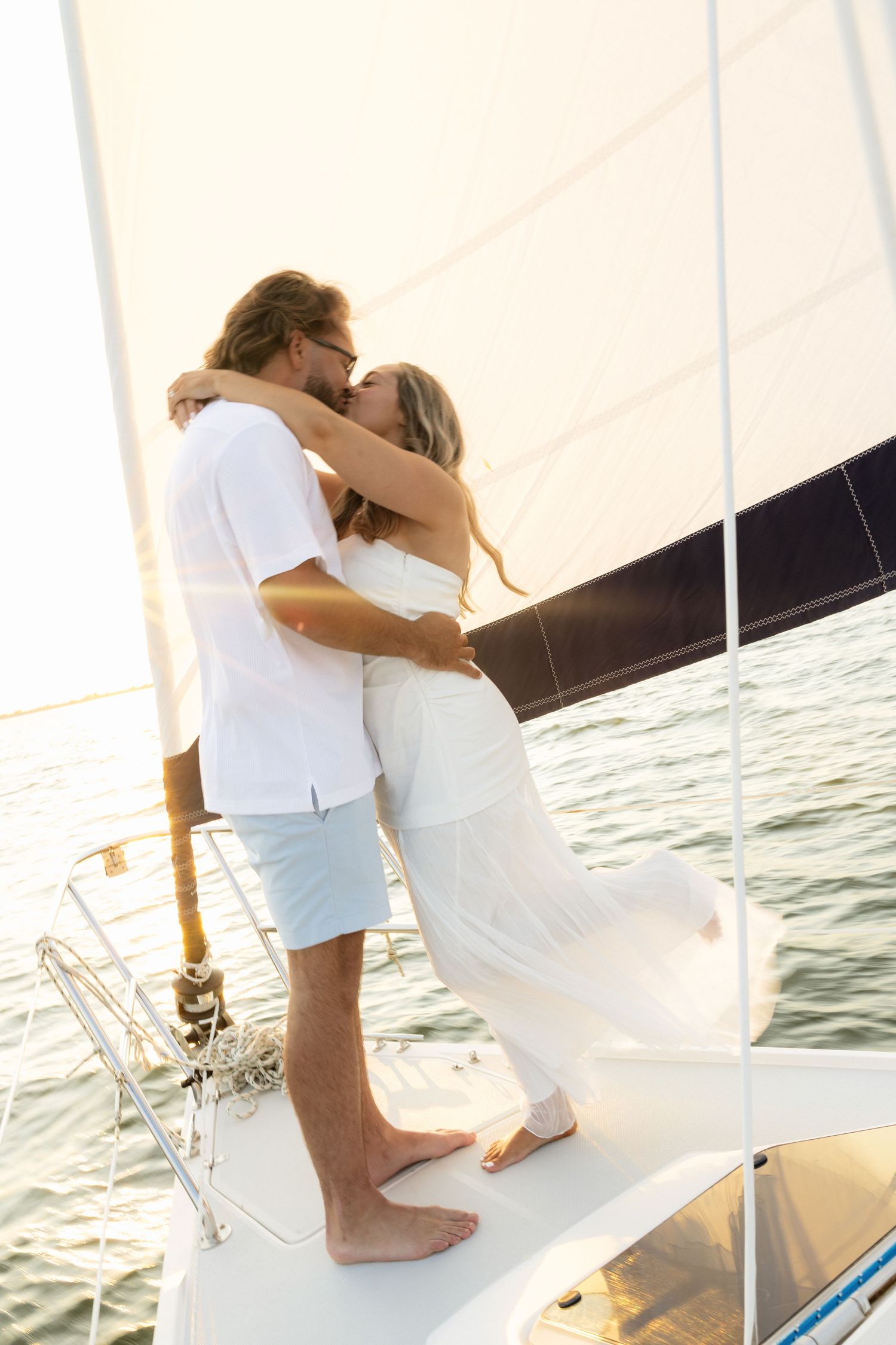 Romantic couple kissing on sailboat deck at sunset, dressed in white, embracing on the bow with ocean views.
