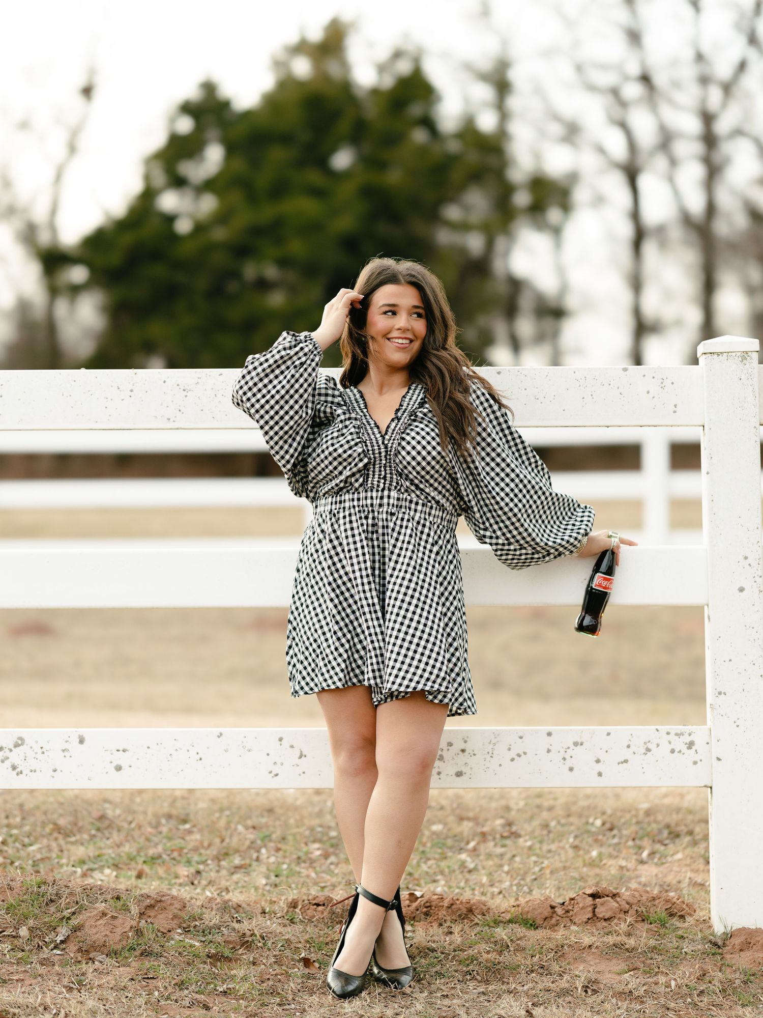 Smiling young woman in houndstooth dress holding a Coca-Cola bottle leaning on a white fence.