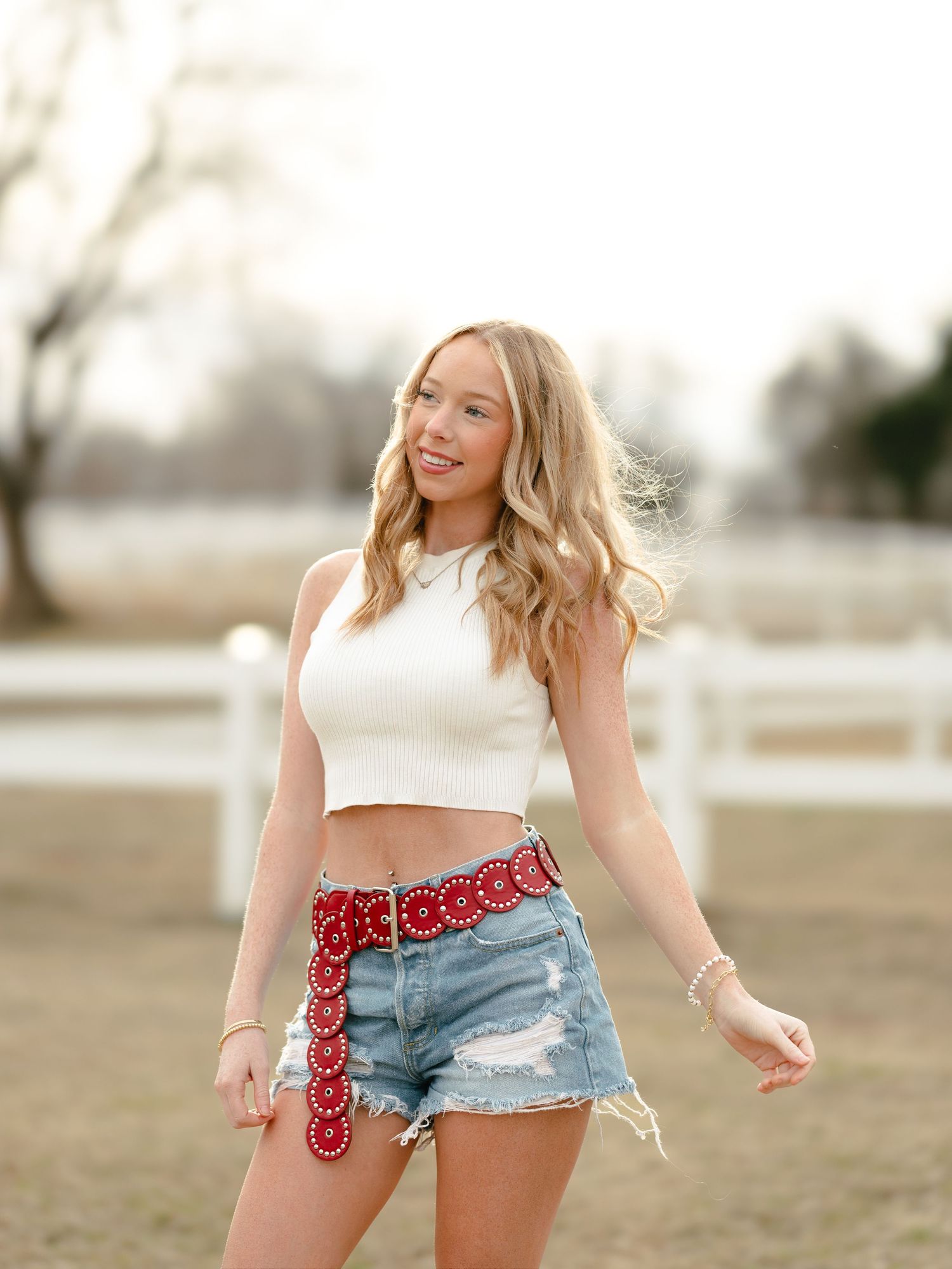 Blonde teen in white crop top and denim cutoffs with red belt posing at ranch.
