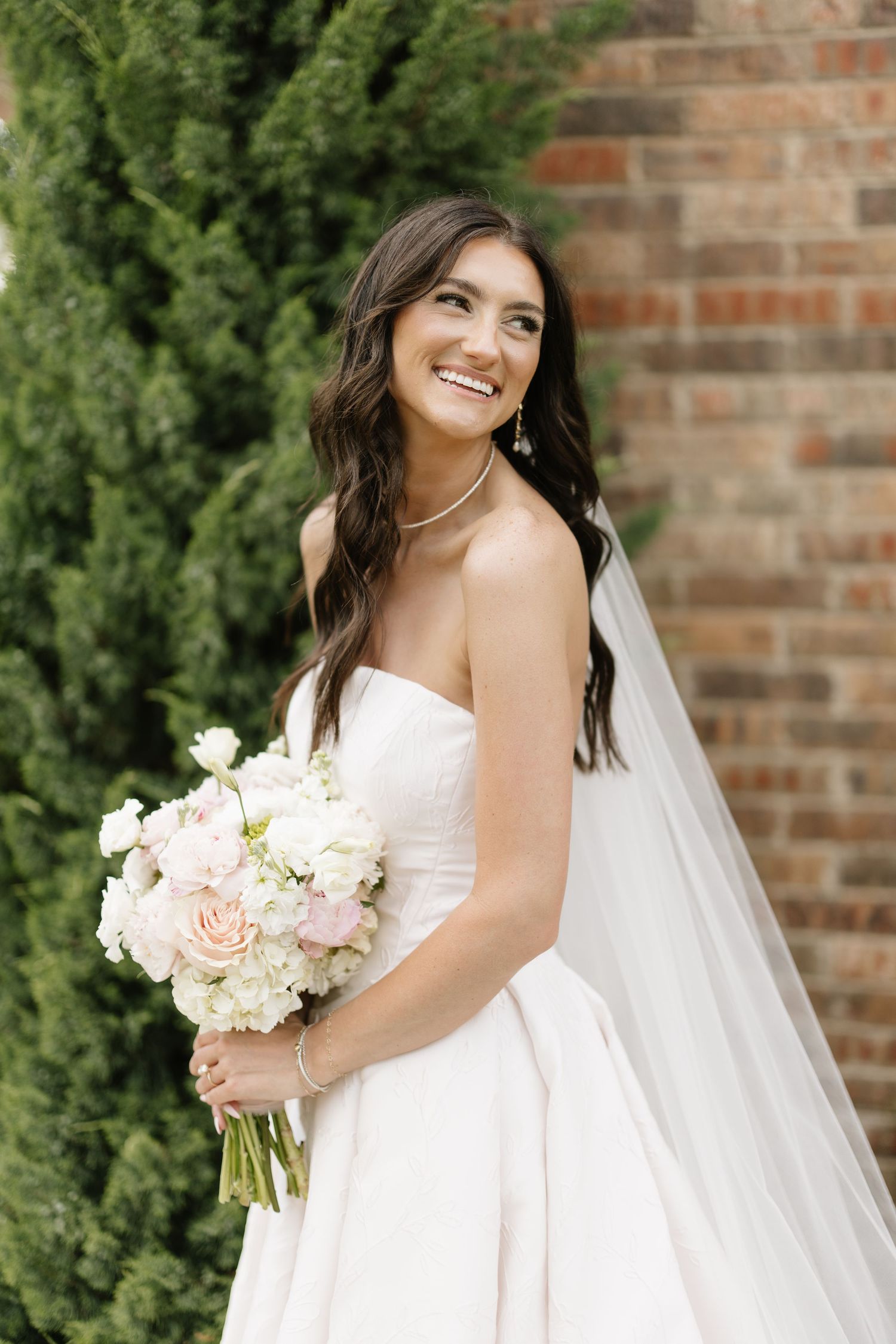 Smiling bride in strapless white ballgown holding pink and white bouquet beside brick wall and cypress trees.