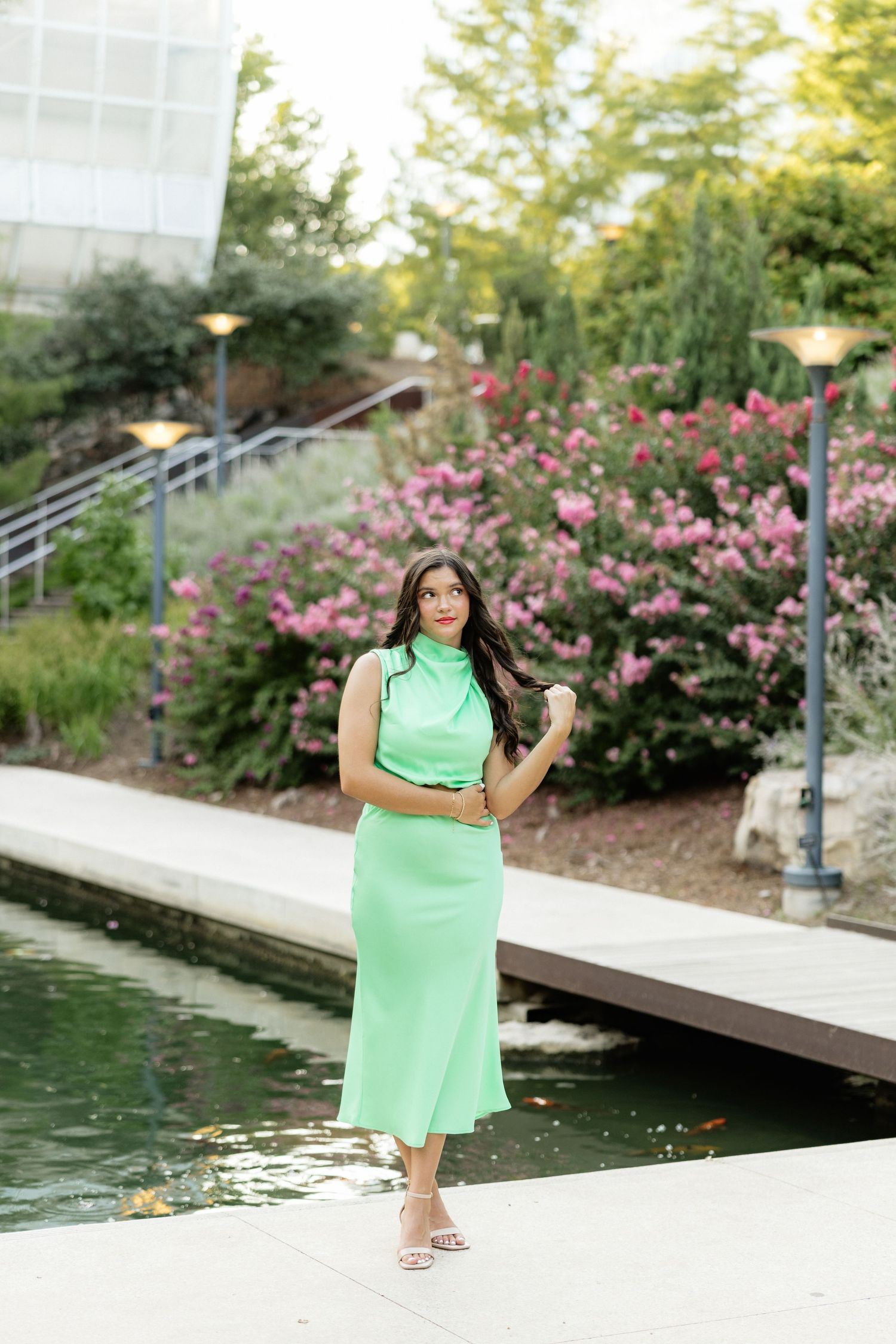 Woman in mint green midi dress posing near a pond with pink flowering bushes in a garden setting.