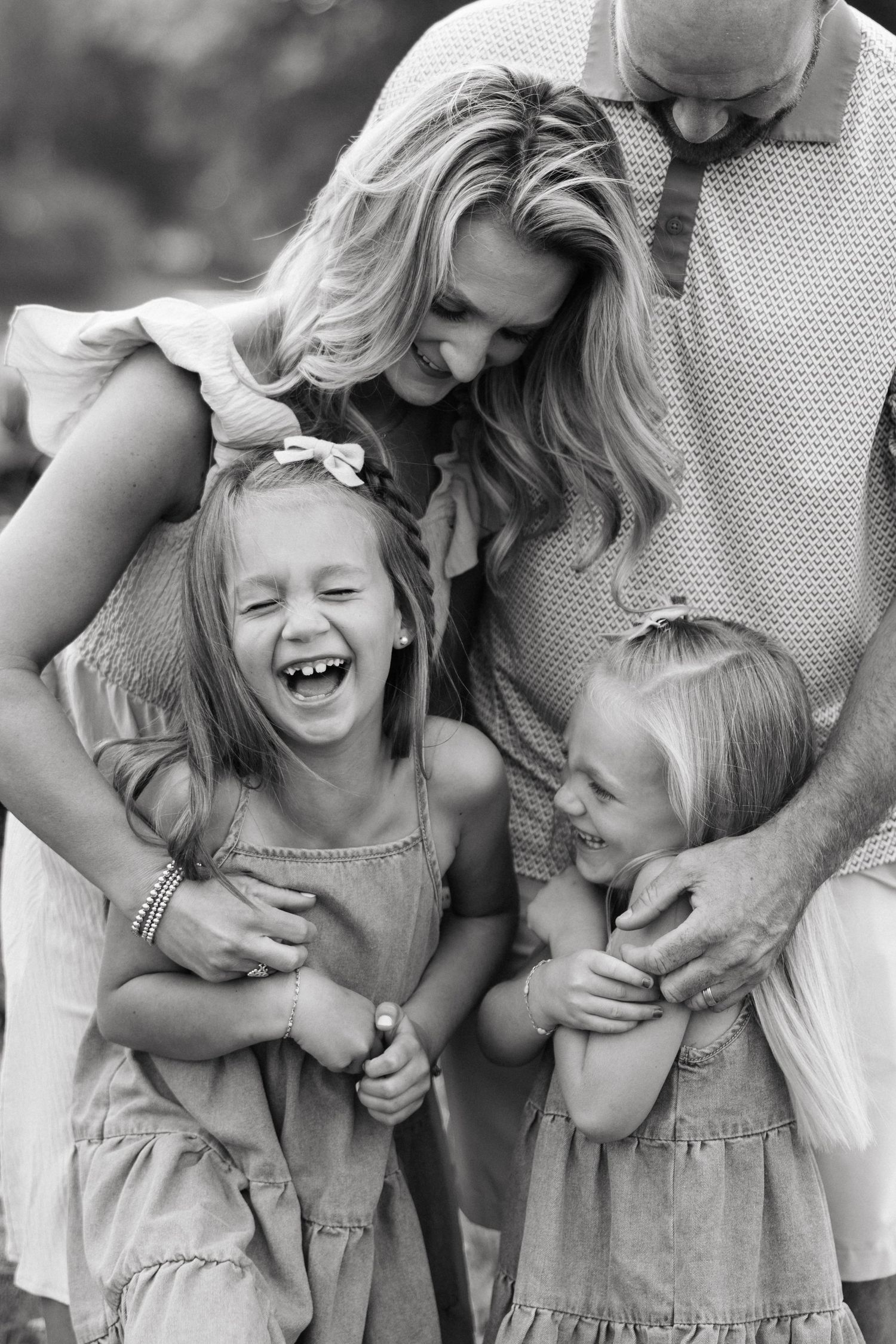 Black and white photo of a joyful family with two laughing blonde girls being hugged by their smiling parents outdoors.