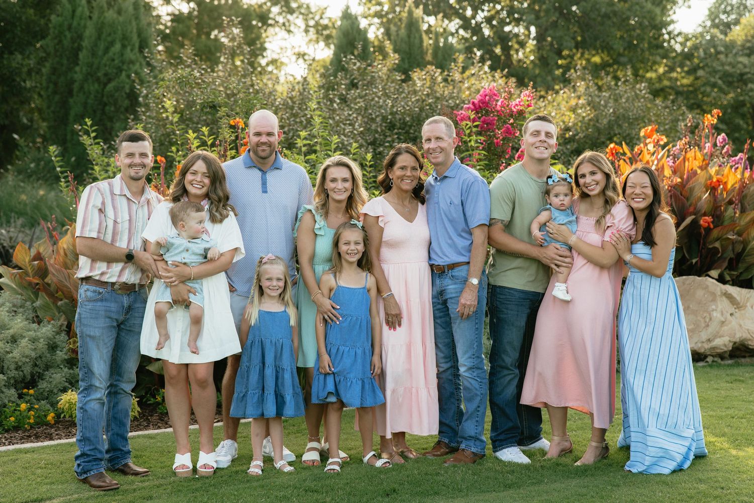 Large family group portrait smiling outdoors in a lush garden setting, wearing colorful summer outfits.
