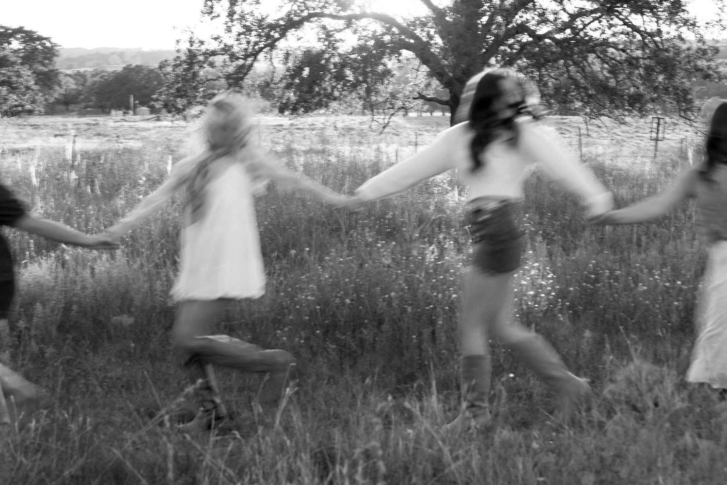 Black and white photo of group of women running and holding hands through a wildflower meadow near trees.