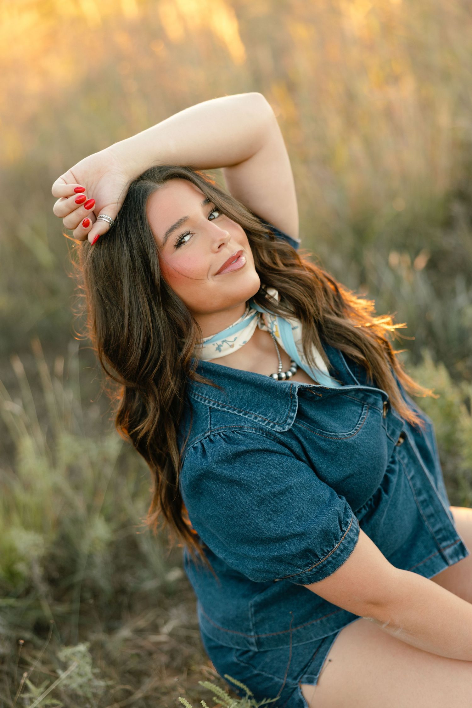 Brunette teen girl in denim romper posing gracefully in a golden field at sunset.