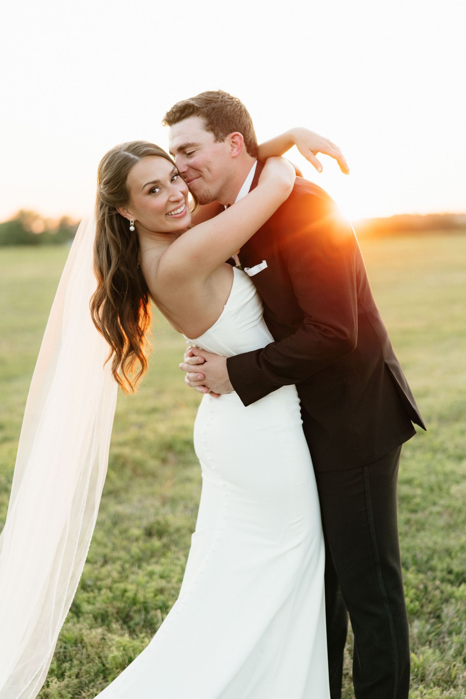 Bride and groom embrace at golden hour sunset in an open field, bride wearing a sleek white gown with veil.