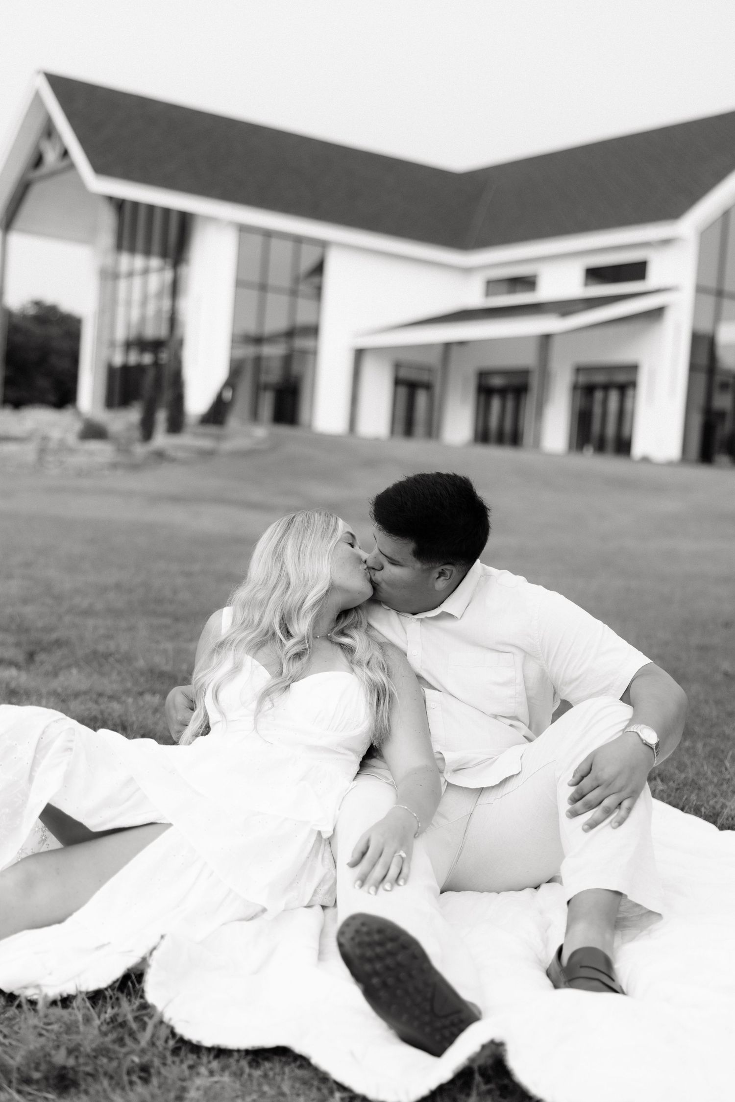 Black and white photo of a couple kissing while sitting on a blanket on grass in front of a white building.