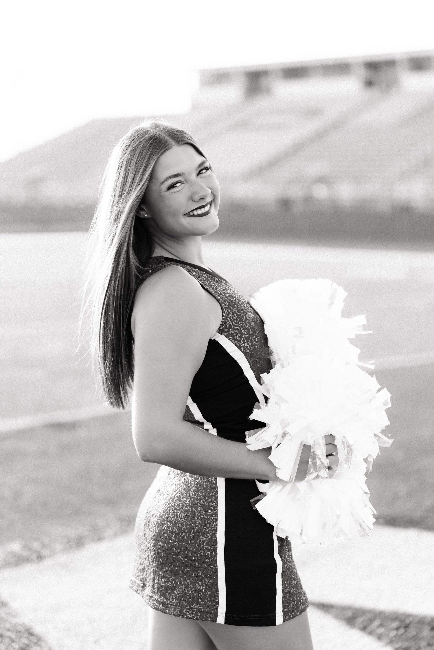 Black and white photo of a smiling cheerleader in uniform holding pom poms at a football stadium.