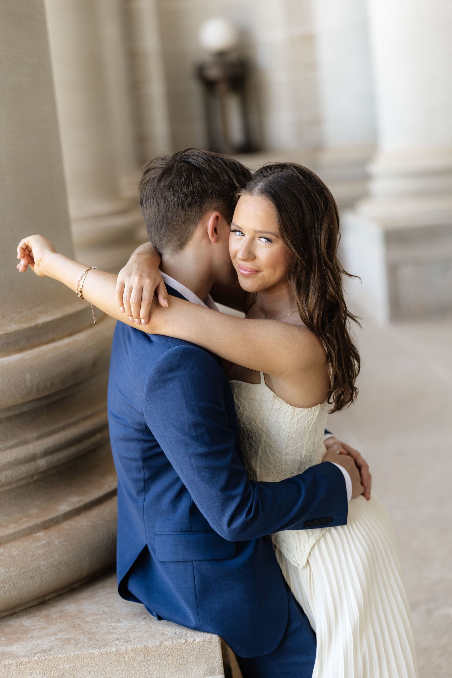A bride in a white dress embraces a groom in a blue suit near elegant stone columns in a romantic wedding portrait.