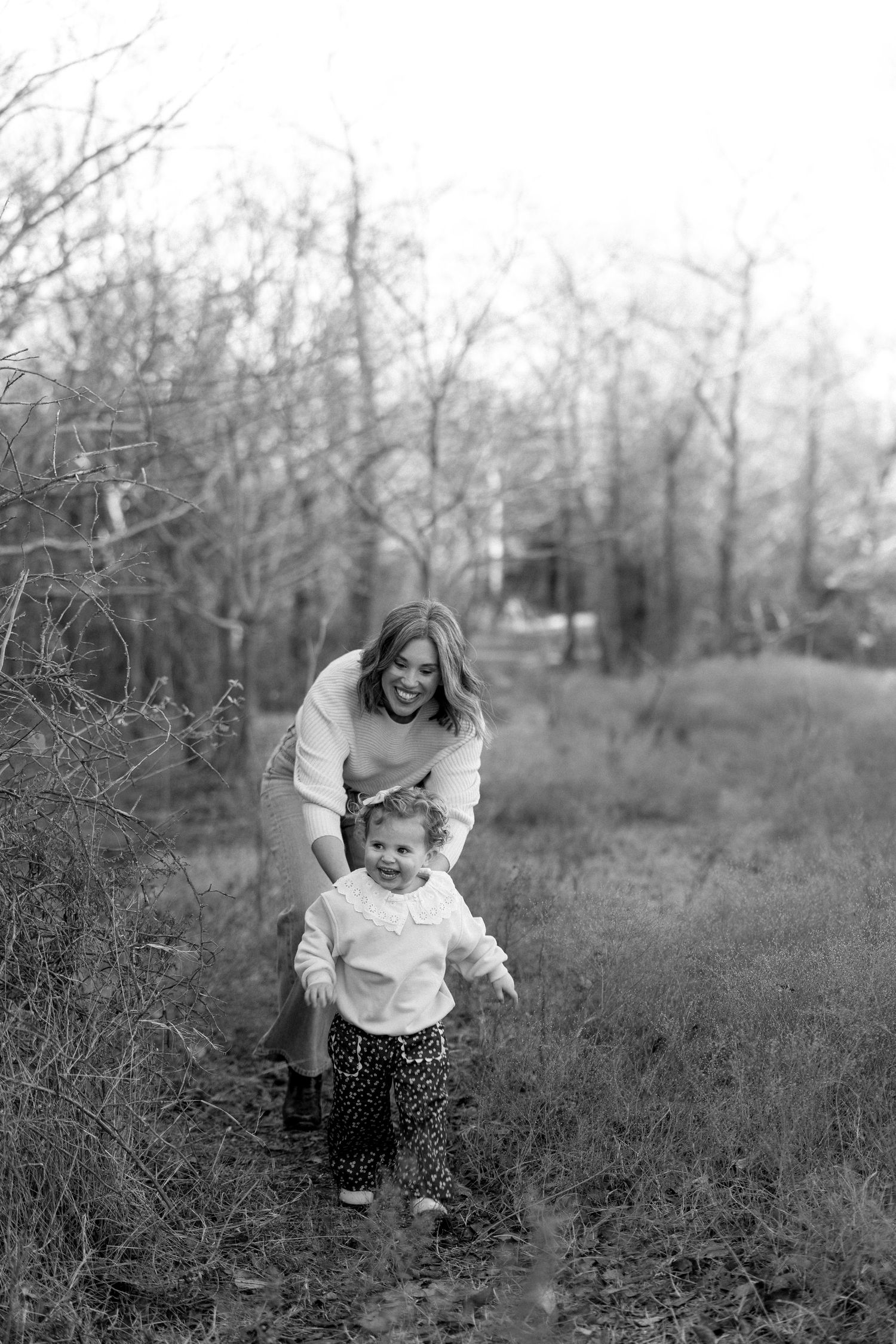 Black and white photo of a smiling mother helping her toddler walk through a wooded outdoor setting.