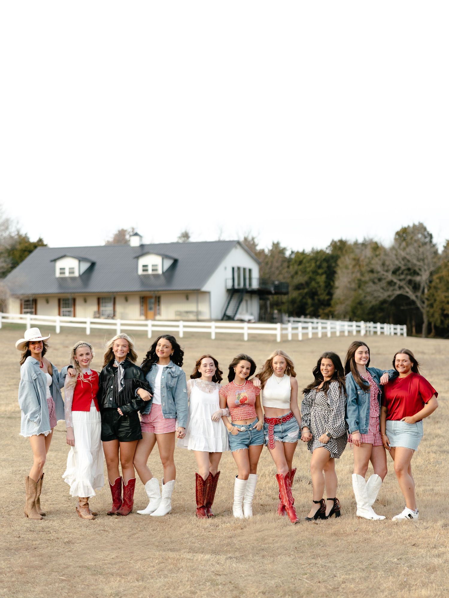Group of ten young women in colorful outfits and cowboy boots posing on a farm with white fence.