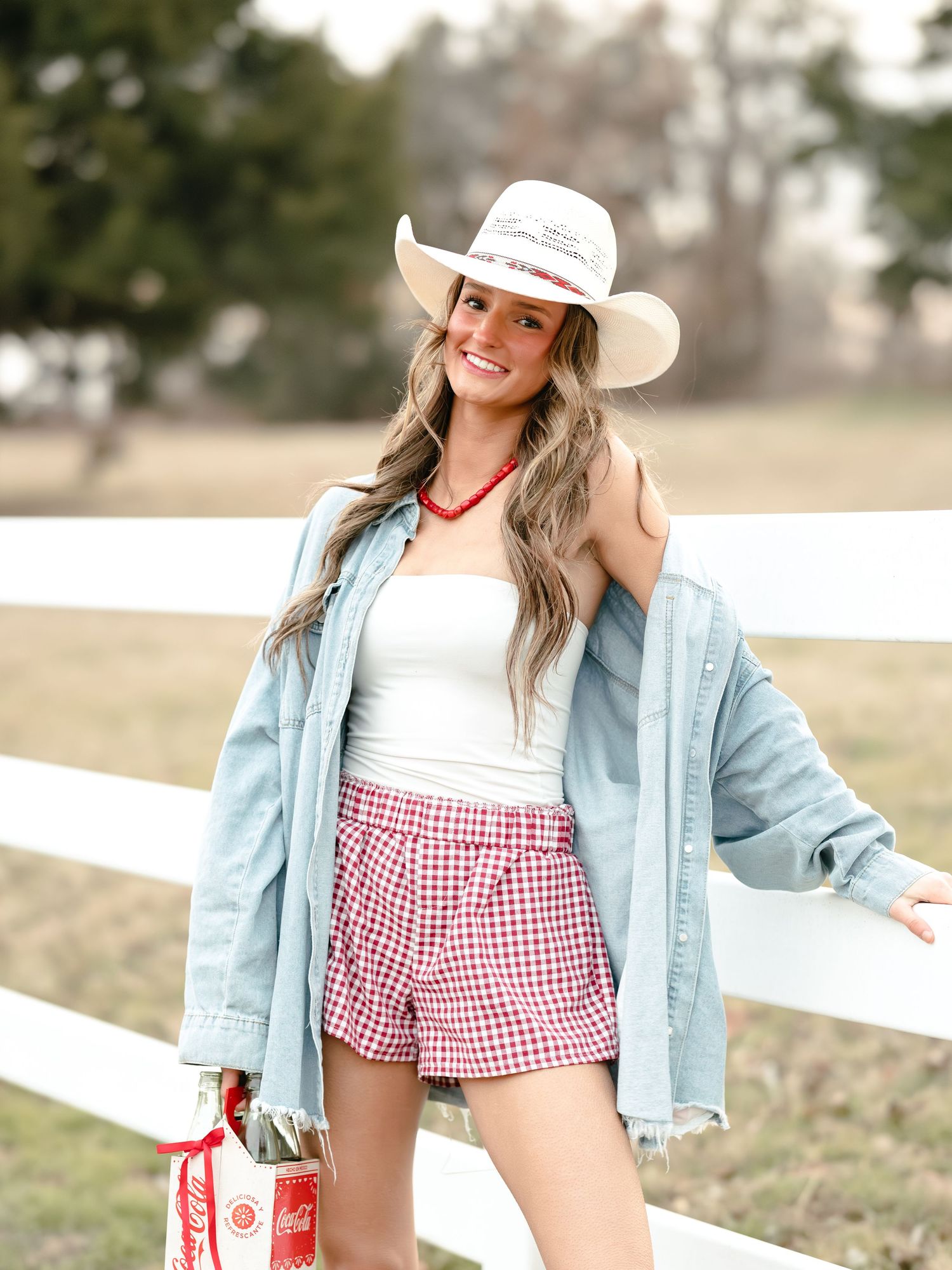 Smiling woman in white tube top, denim shirt, red gingham shorts, and cowboy hat by white fence.