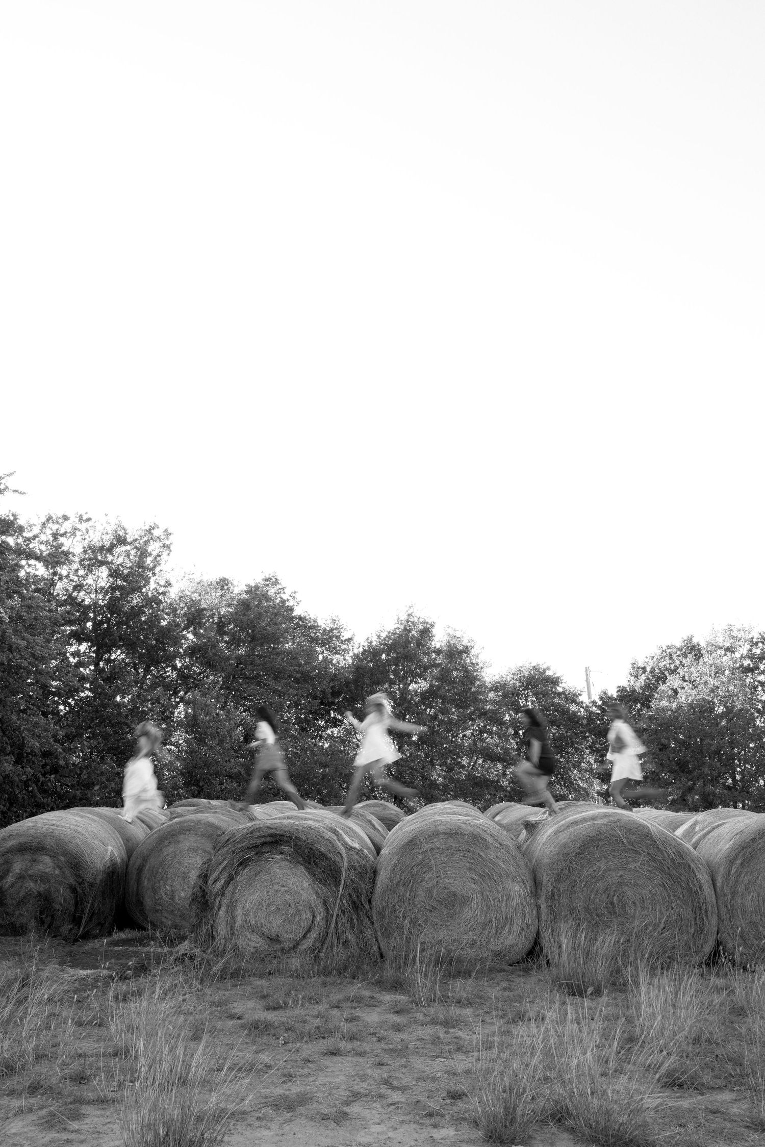 Black and white photo of girls jumping and running across large hay bales outdoors.