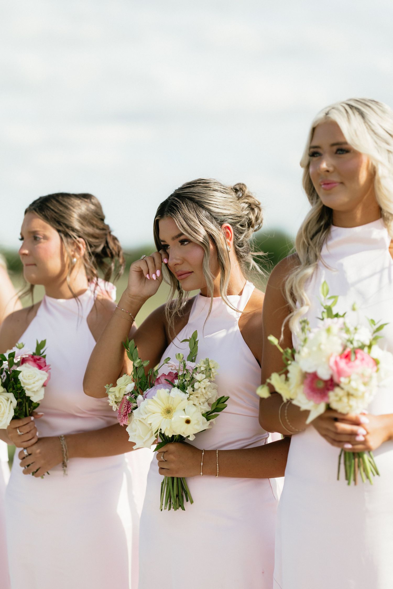 Three bridesmaids in blush pink dresses holding white and pink floral bouquets at an outdoor wedding ceremony.