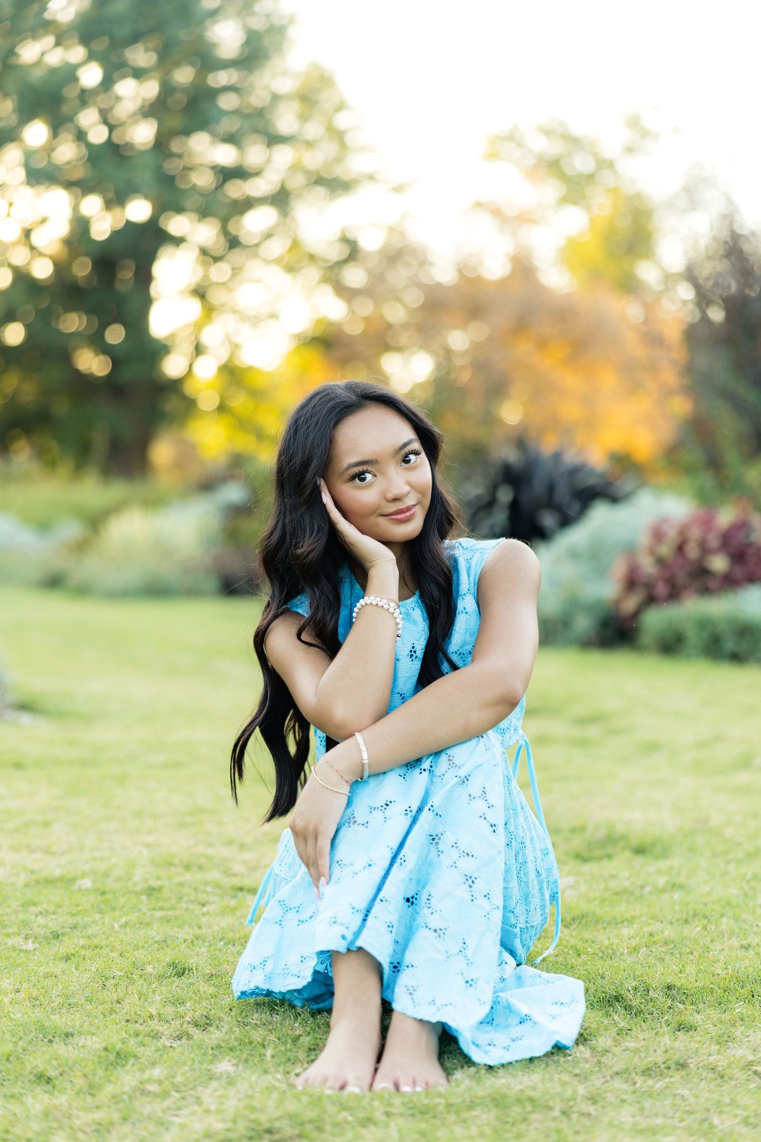 Young woman in light blue floral dress sitting barefoot on grass in a garden at golden hour.