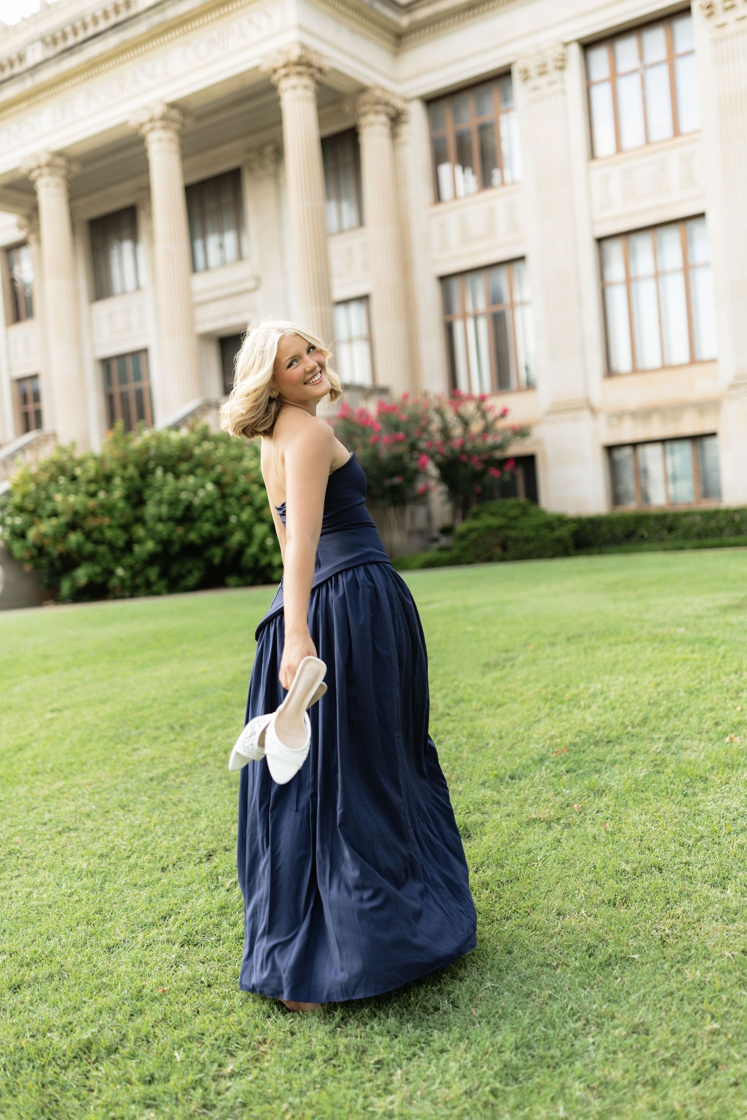 Smiling blonde woman in navy blue strapless ball gown holding white heels on green lawn before grand building.