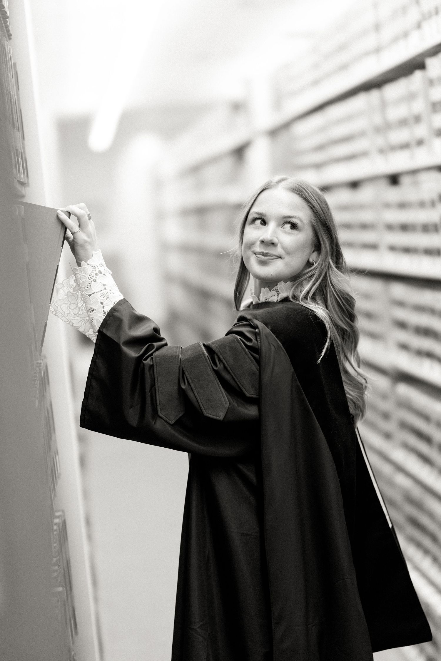 Graduate in black gown writing on whiteboard in library, black and white.