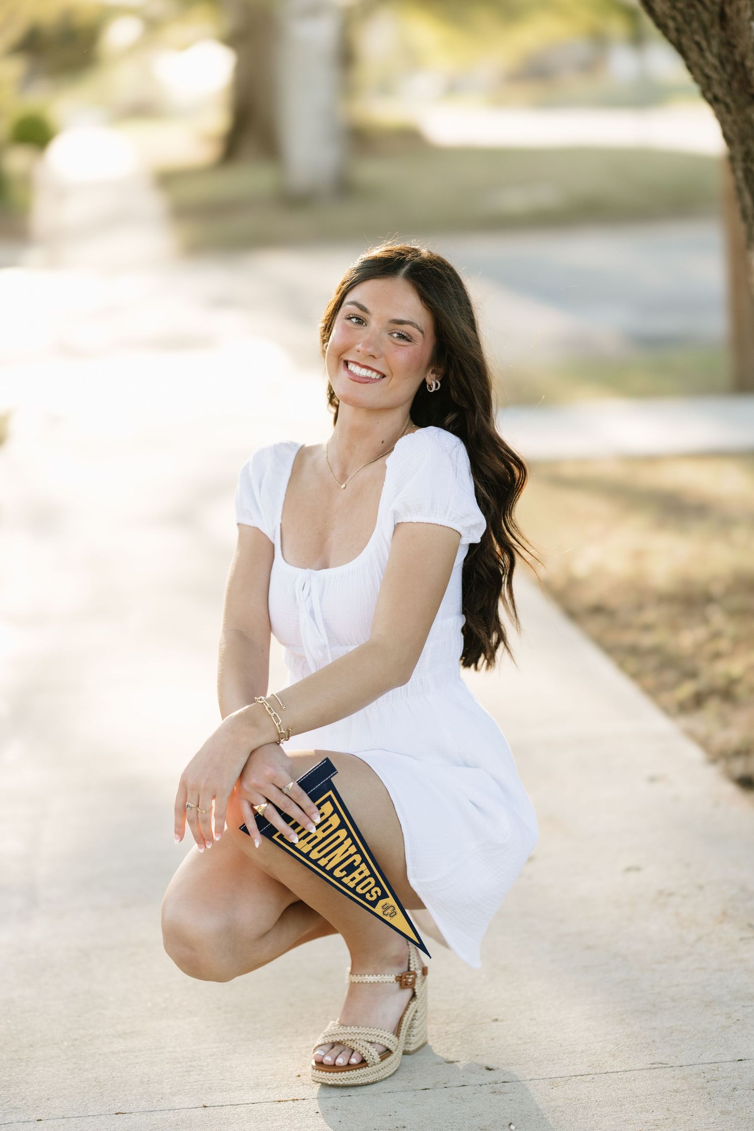 Smiling brunette woman in white dress crouching on sidewalk holding a college pennant flag.