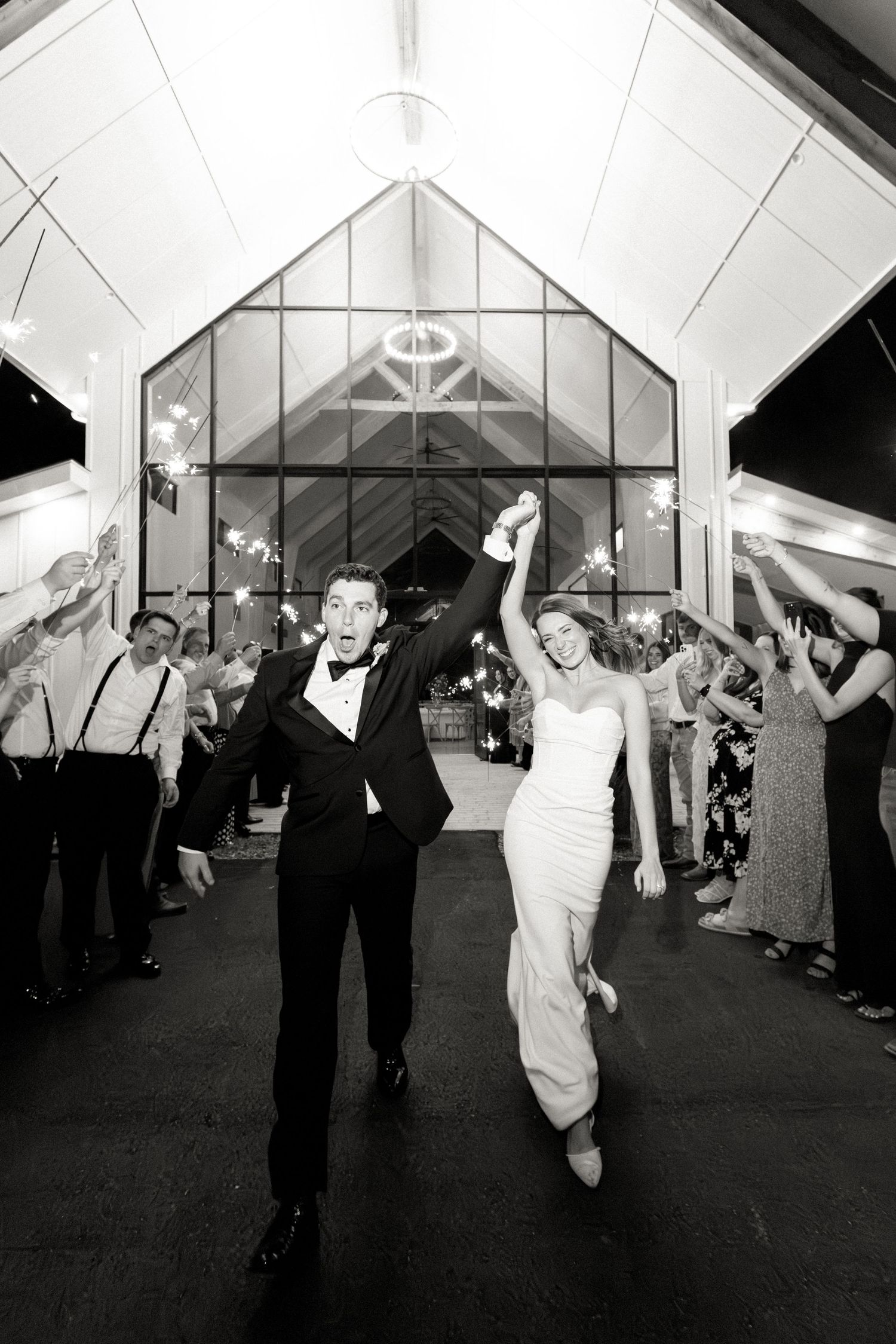 Black and white photo of bride and groom exiting through sparkler sendoff in elegant glass chapel.