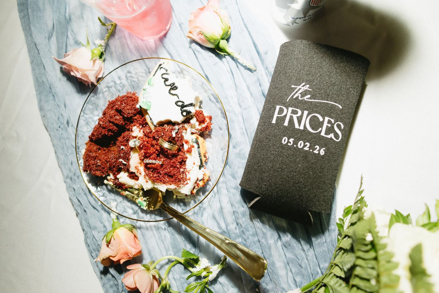 Wedding details flat lay featuring red velvet cake, personalized koozie reading The Prices 05.02.26, and pink roses.