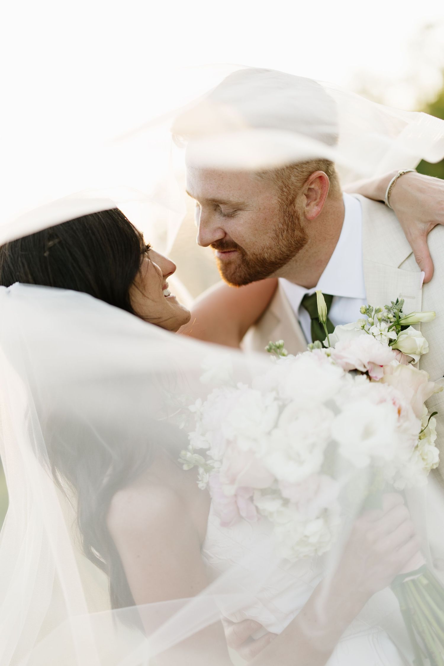 Romantic bride and groom gazing at each other wrapped in a flowing veil holding white bouquet.