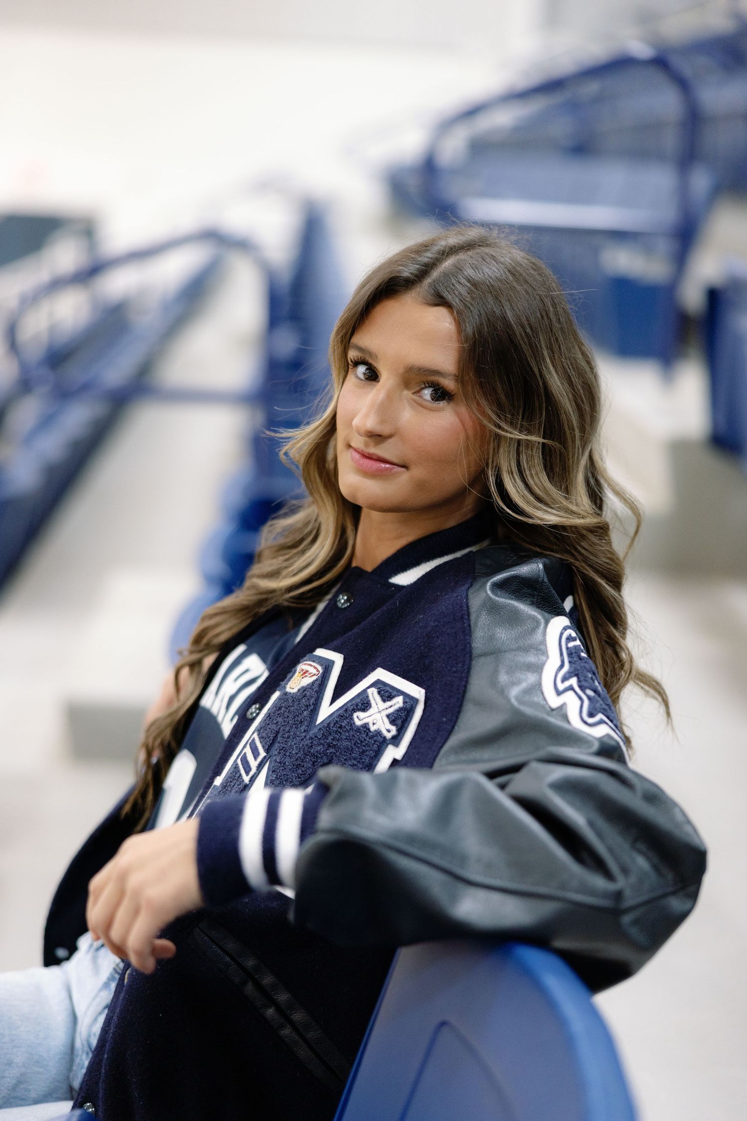 Young woman in varsity jacket poses in stadium bleachers for senior portrait photo.