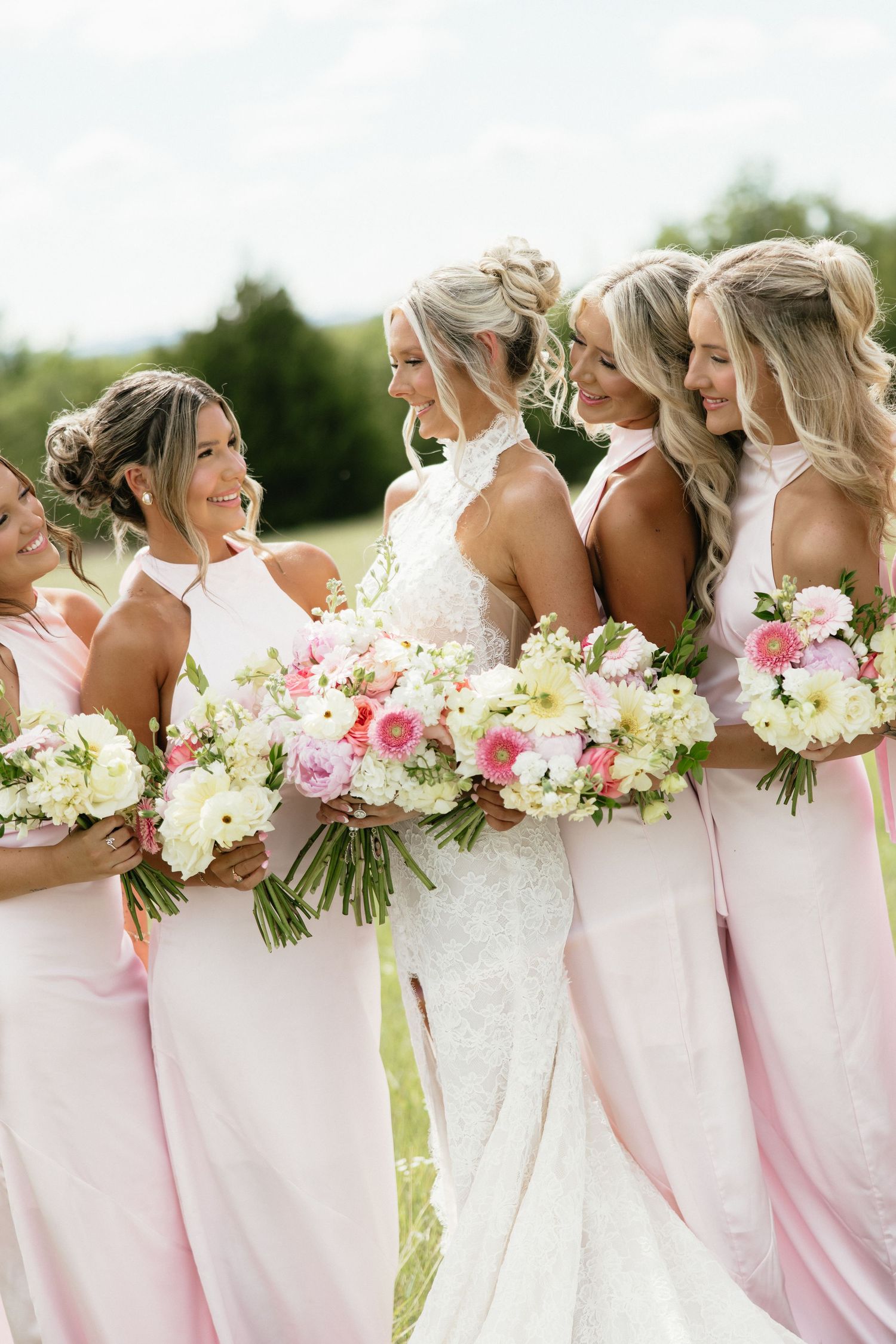 Bride in lace gown poses with bridesmaids in blush pink dresses holding pink and white bouquets.