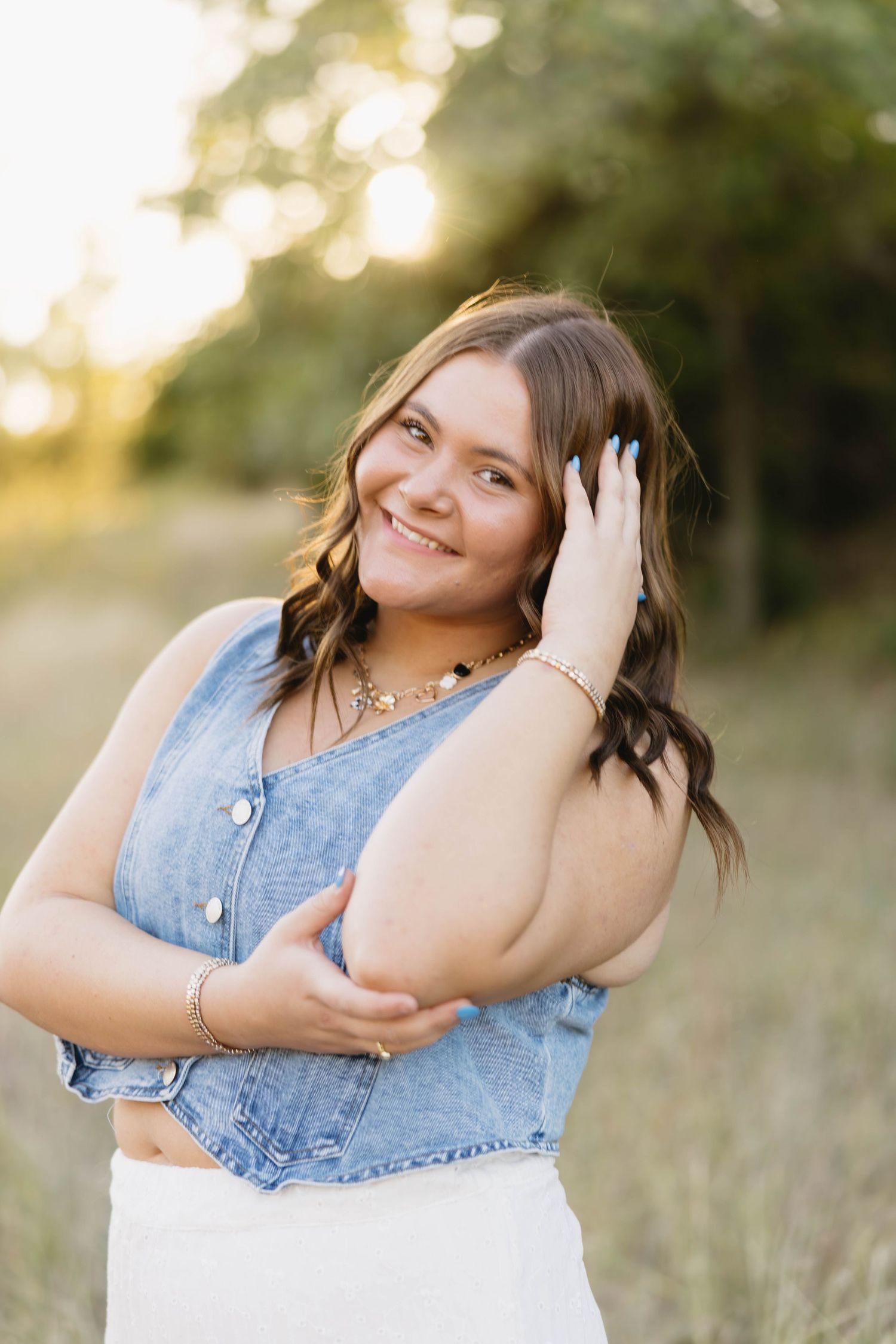 Smiling young woman in denim vest and white skirt posing outdoors at golden hour.