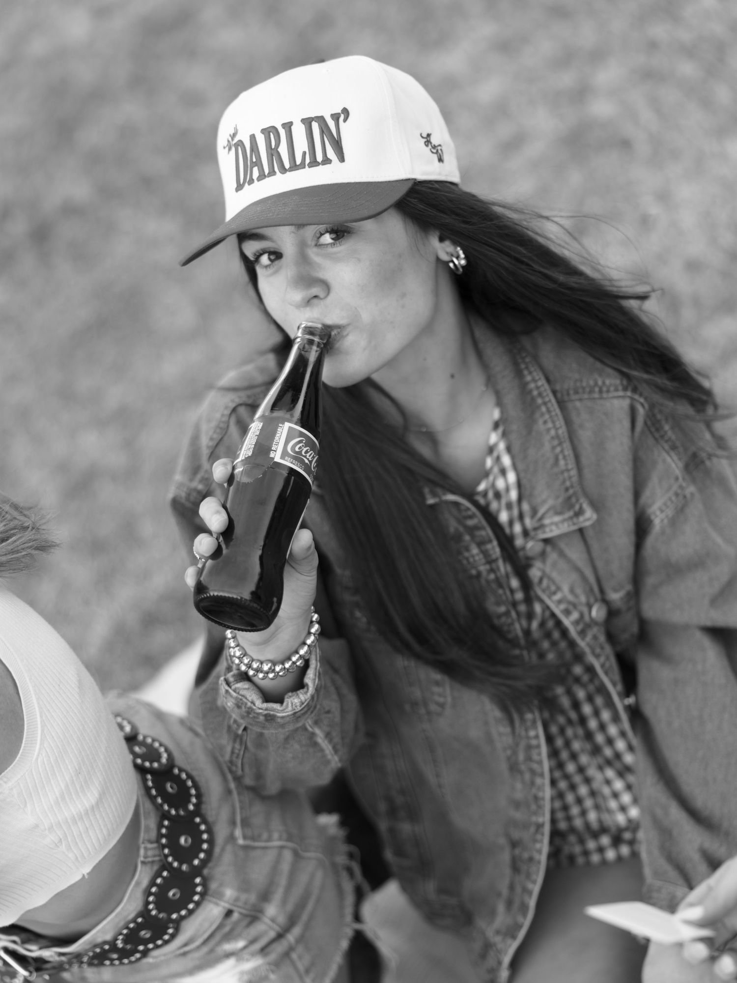 Black and white photo of woman in 'Darlin' cap drinking Coca-Cola bottle at outdoor gathering.