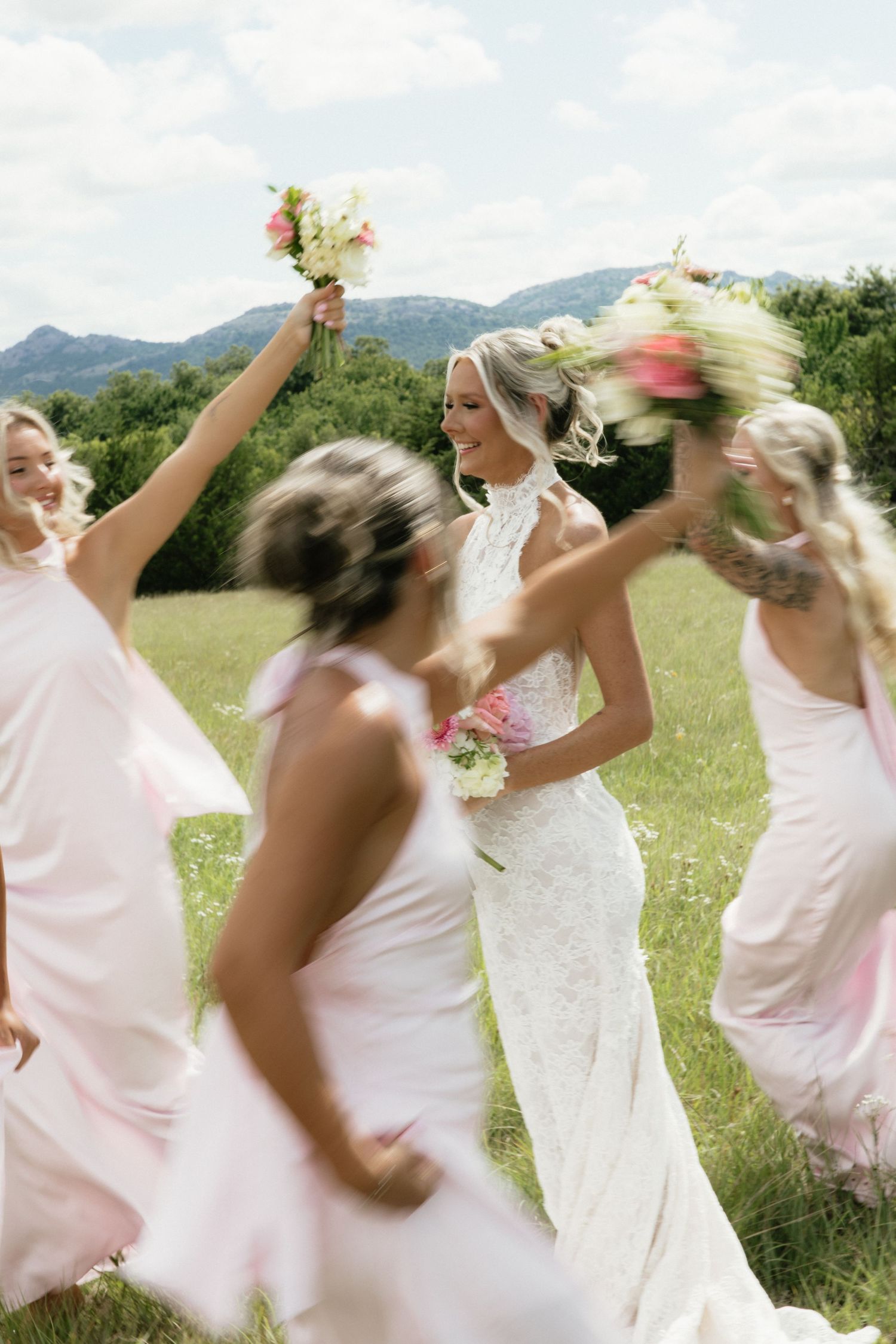 Bride in white lace gown celebrates with bridesmaids in pink dresses, holding bouquets in a sunny green field with mountains.
