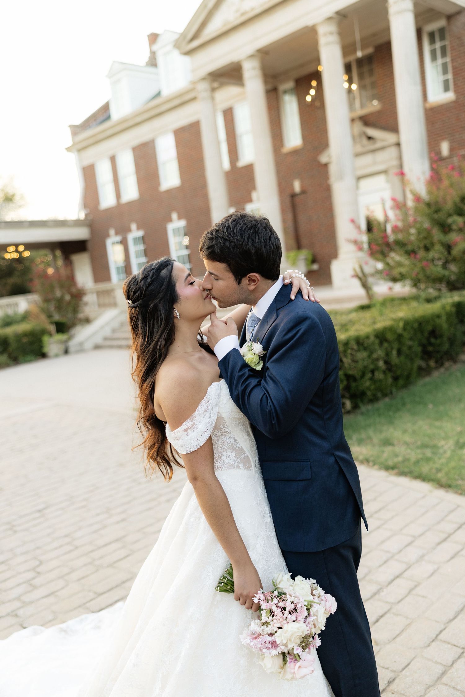 Bride and groom sharing a romantic kiss outside a brick mansion, bride holding a pink bouquet in a white off-shoulder gown.