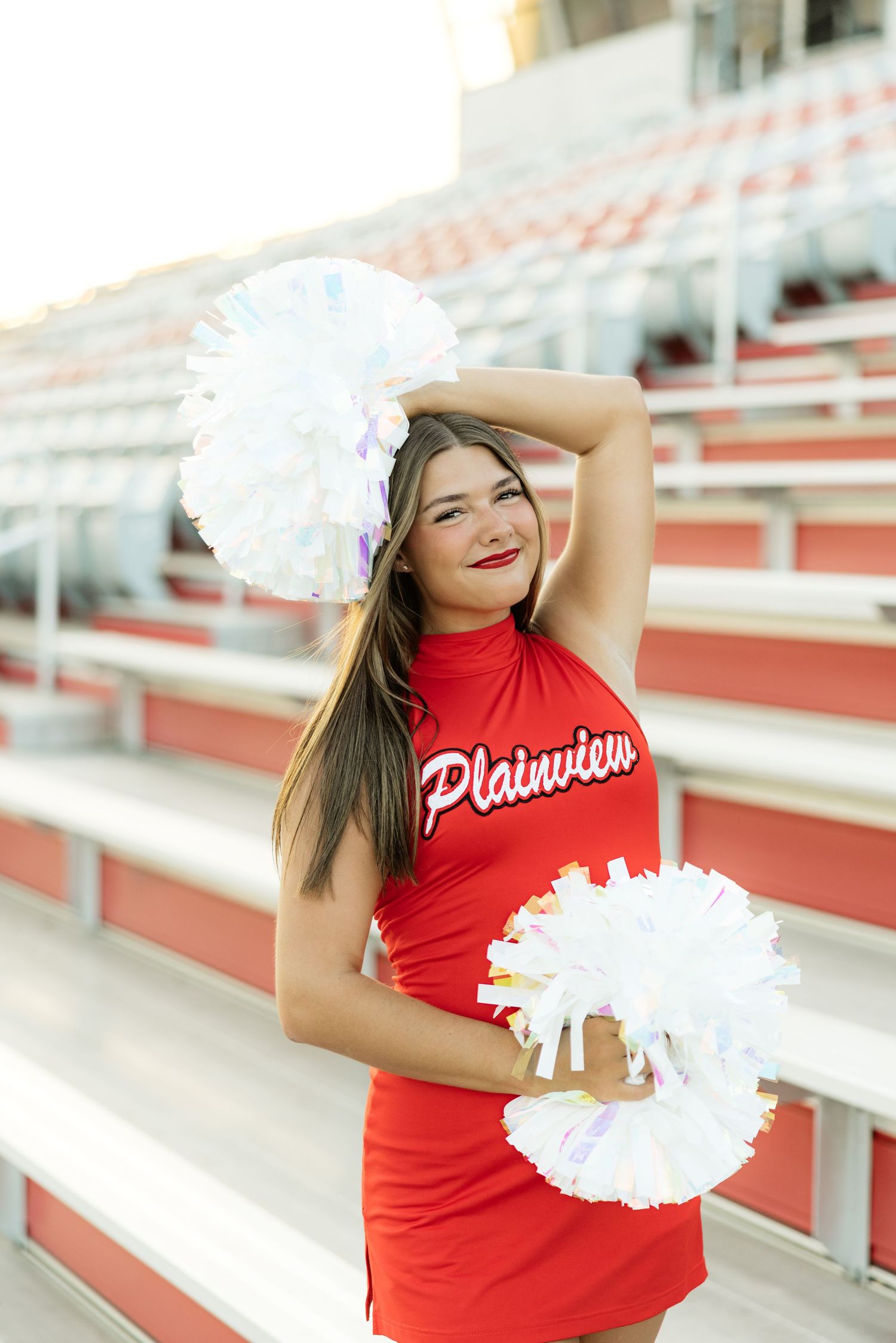 Plainview cheerleader in red uniform poses with white pom-poms in stadium bleachers.